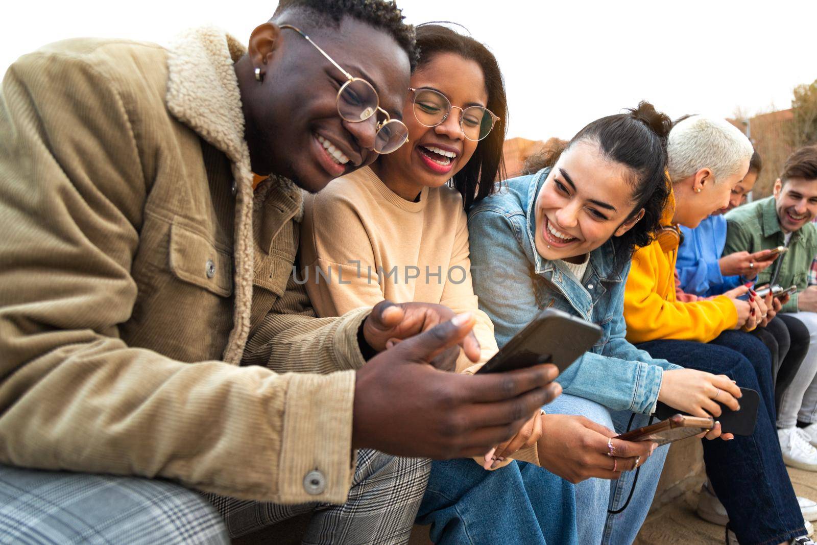 Happy multiracial teen college students having fun using mobile phone together in campus outdoors. Friendship and technology concept.
