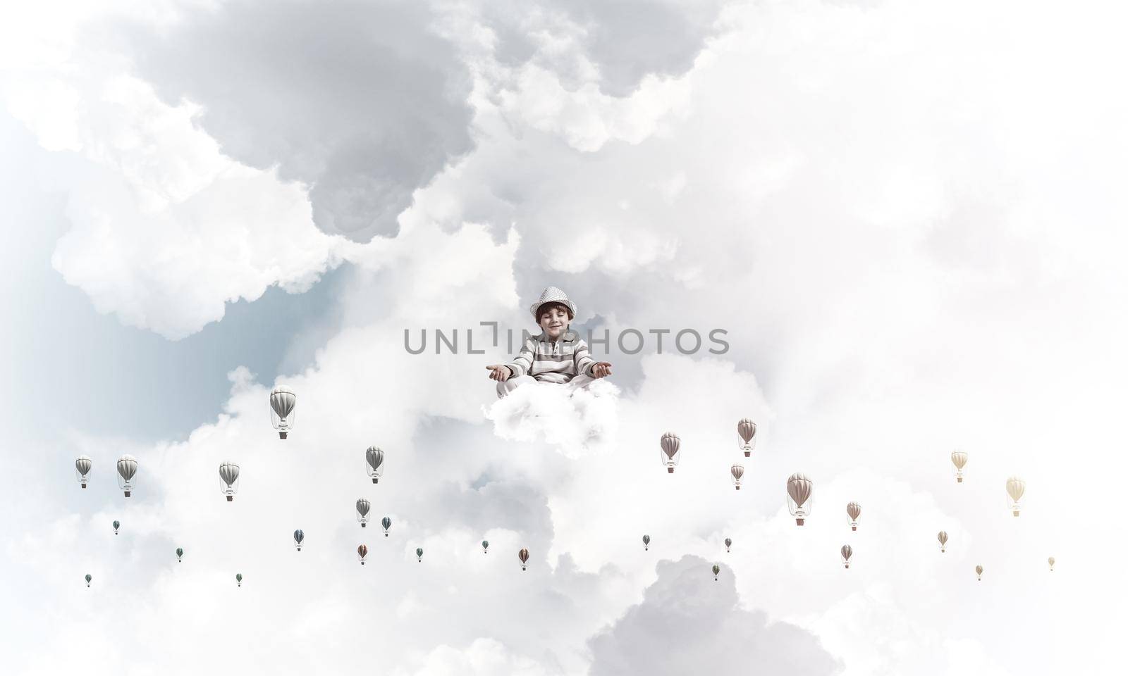 Young little boy keeping eyes closed and looking concentrated while meditating among flying balloons in the air with cloudy skyscape on background.
