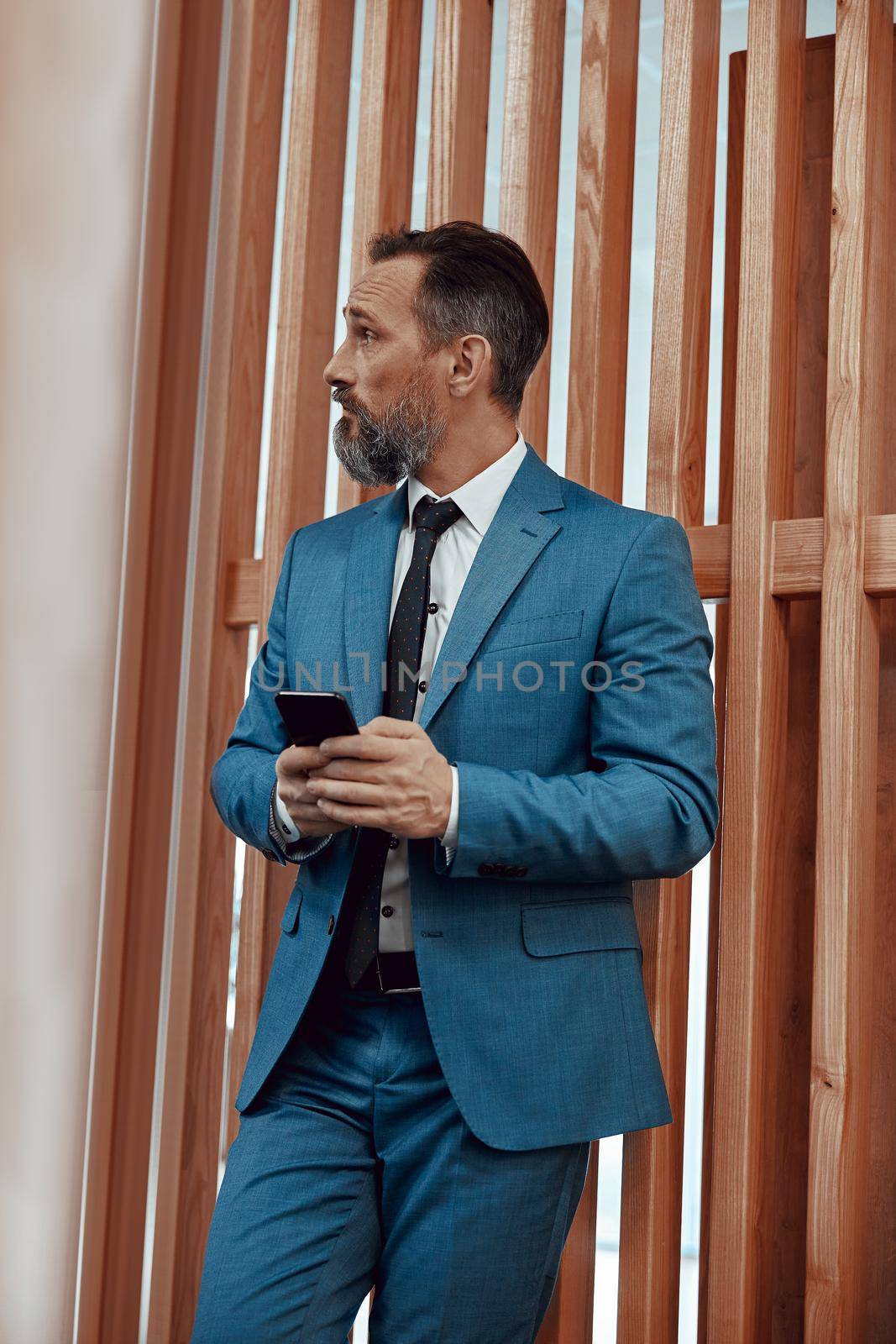 Adult businessman in suit standing and holding mobile phone indoors