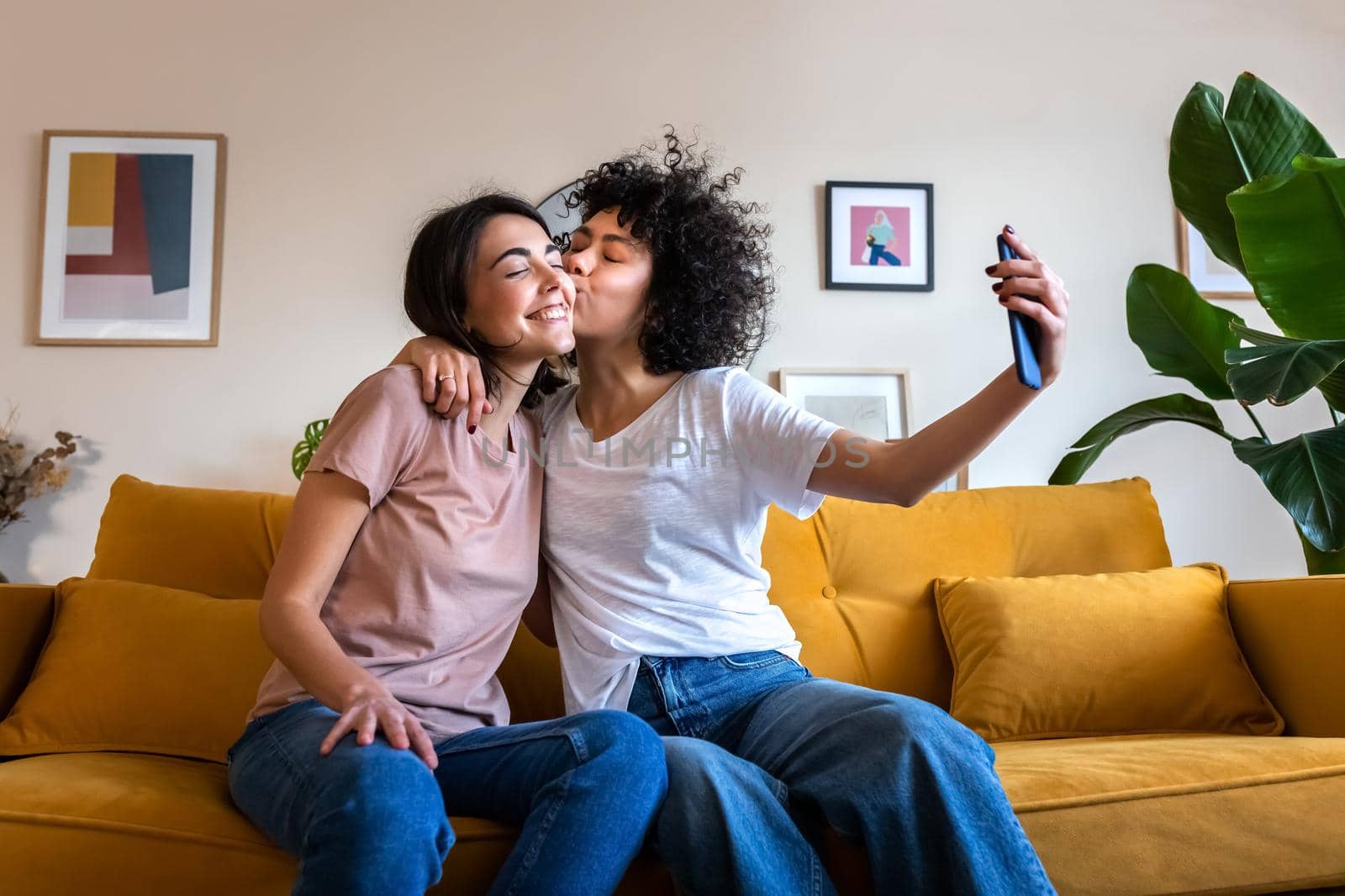Young lesbian couple taking selfie at home using mobile phone. Girlfriend gives kiss on the cheek. by Hoverstock