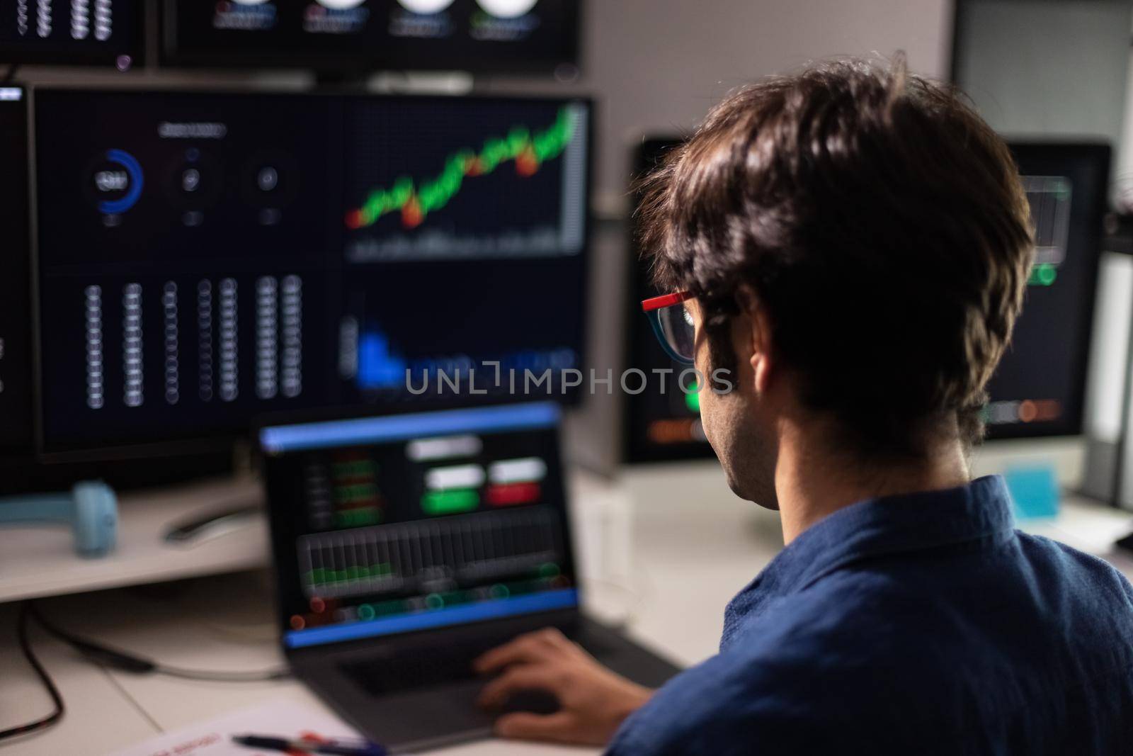 Back view of man in blue shirt working with multiple screens and laptop. Stock market and trading. Business concept.