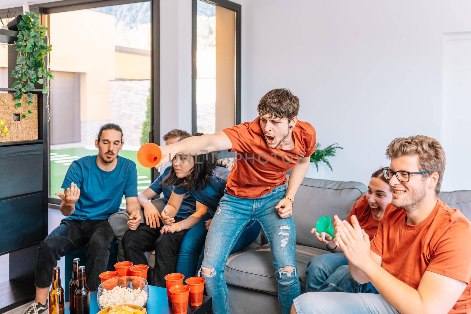 two groups of young friends facing each other watching a sports championship broadcast on TV. in the living room. by CatPhotography