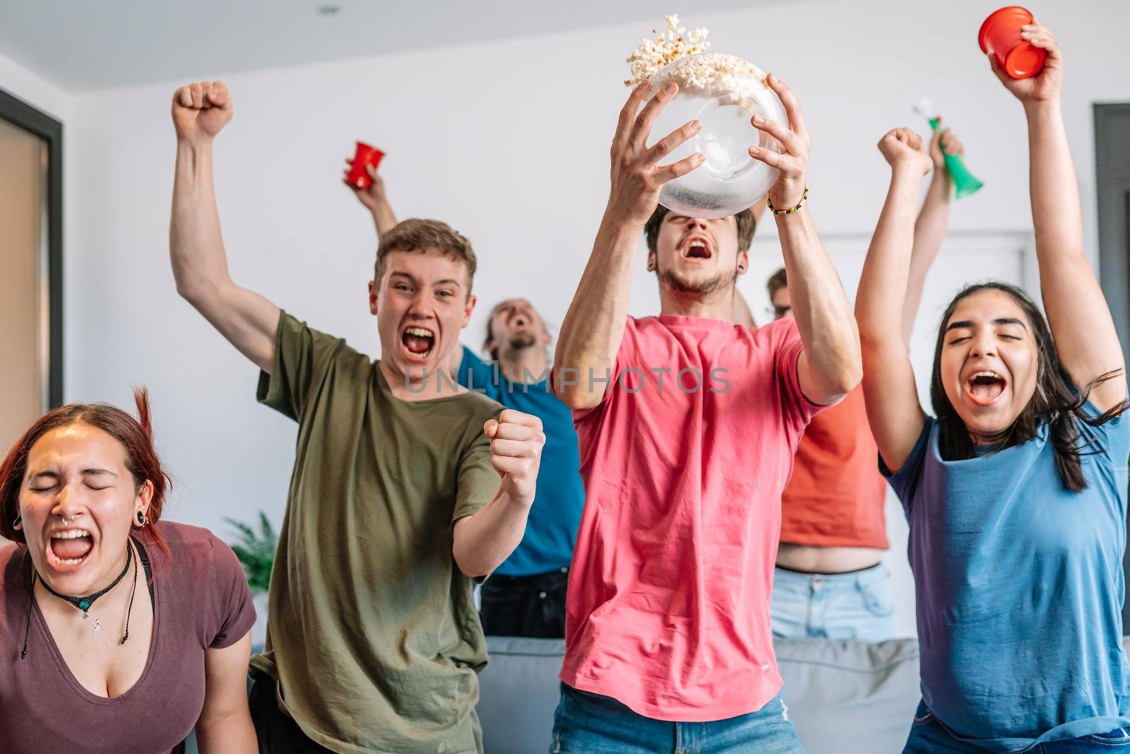 friends jumping for joy, throwing popcorn in the air after their team's victory. group of youngsters partying at home. by CatPhotography