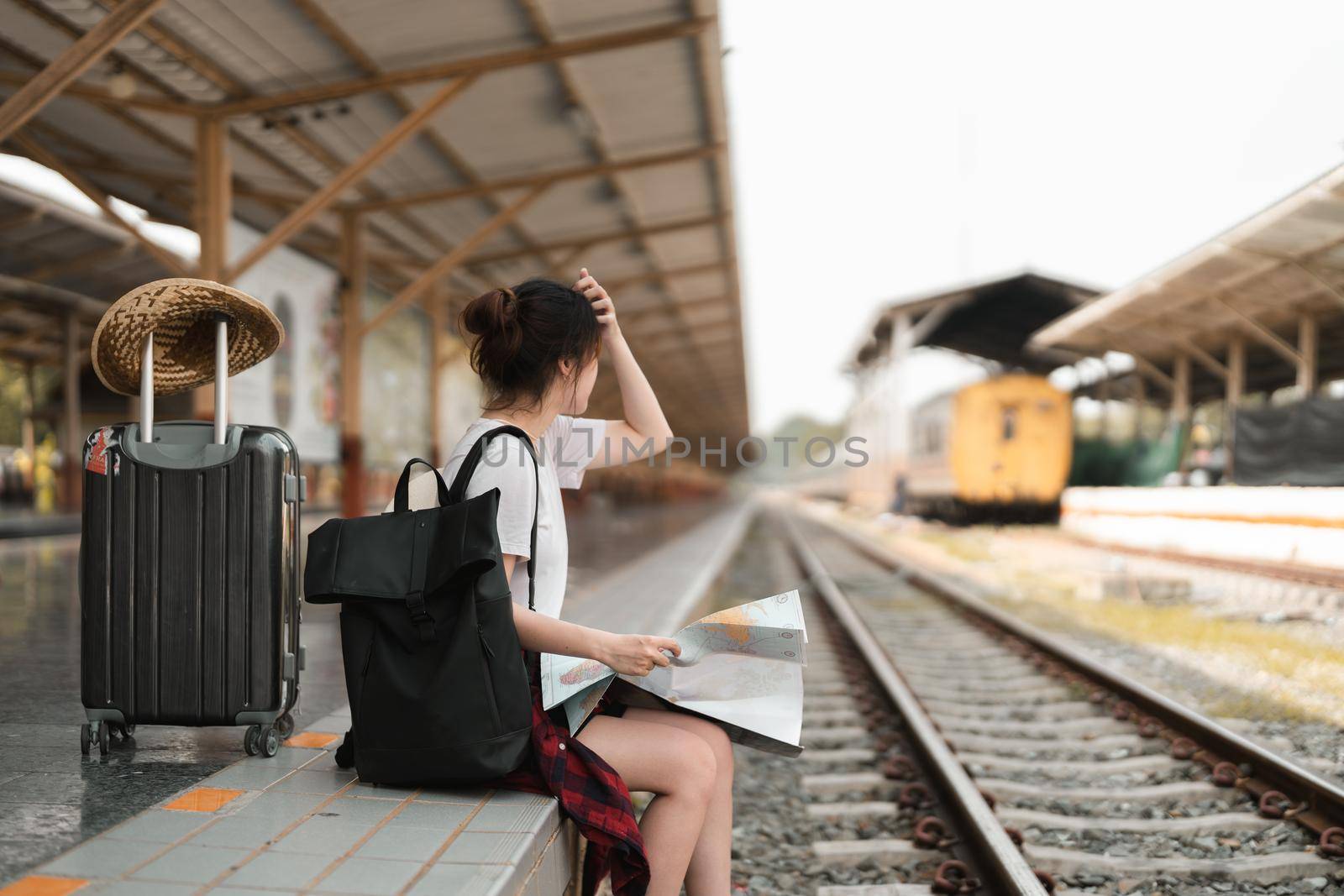 Young woman traveler with backpack looking to map while waiting for train, Asian backpacker on railway platform at train station. Holiday, journey, trip and summer summer travel concept by nateemee