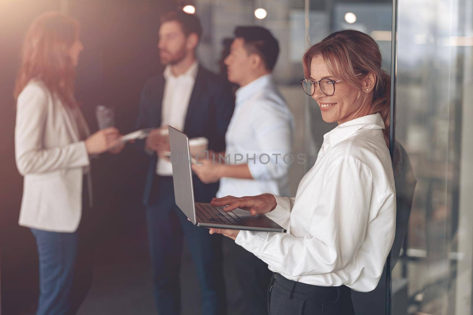 Mature smiling businesswoman at office with group of colleagues on background, working on laptop