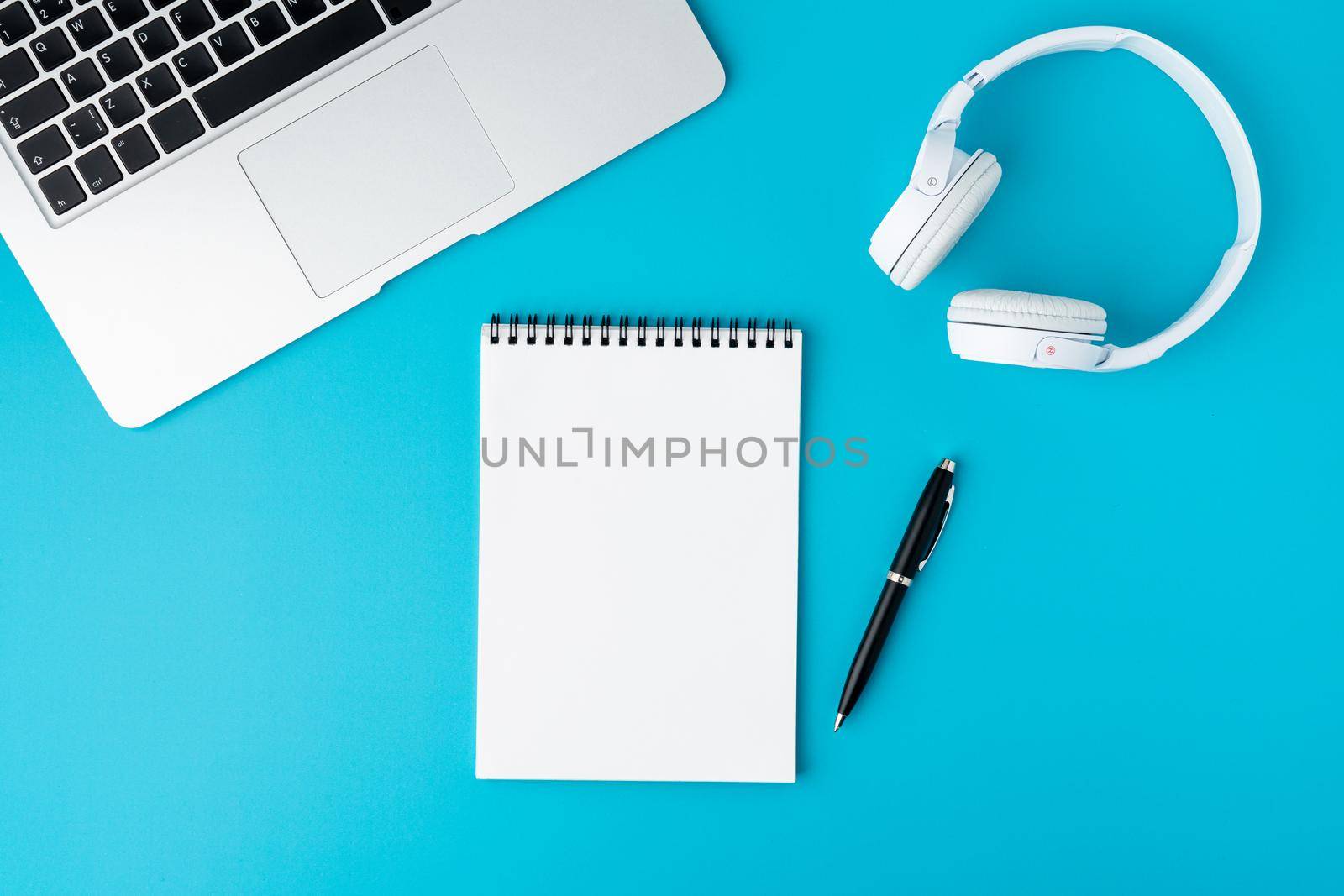 Blank empty spiral notebook on bright blue background. Laptop keyboard, headphones and automatic pen. Concept of modern workplace flat lay. Portable office equipment. Wireless technology, horizontal