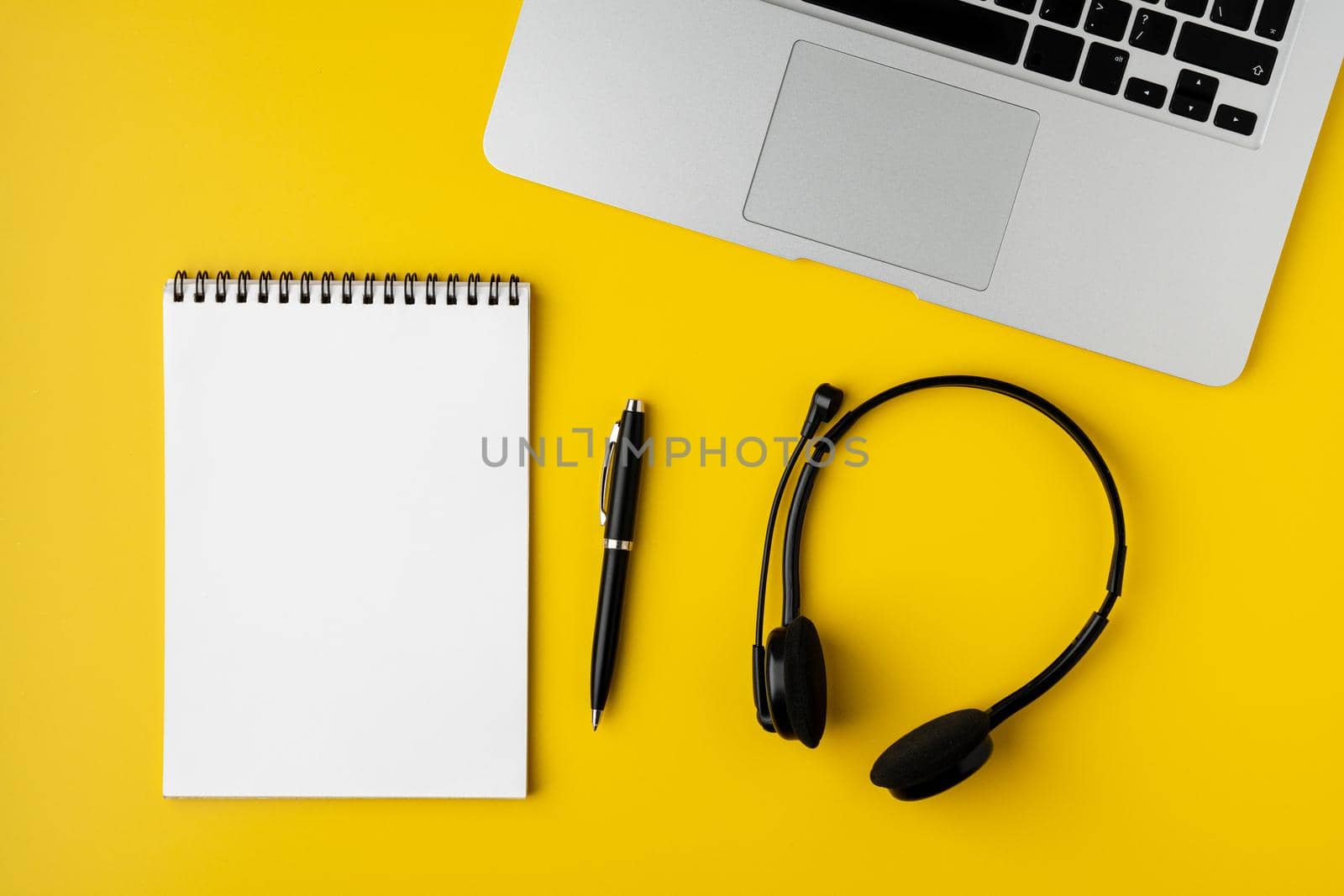 Metallic laptop keyboard, black wireless headset with mic, spiral empty notebook and pen on yellow background. Concept of modern workspace, overhead, horizontal