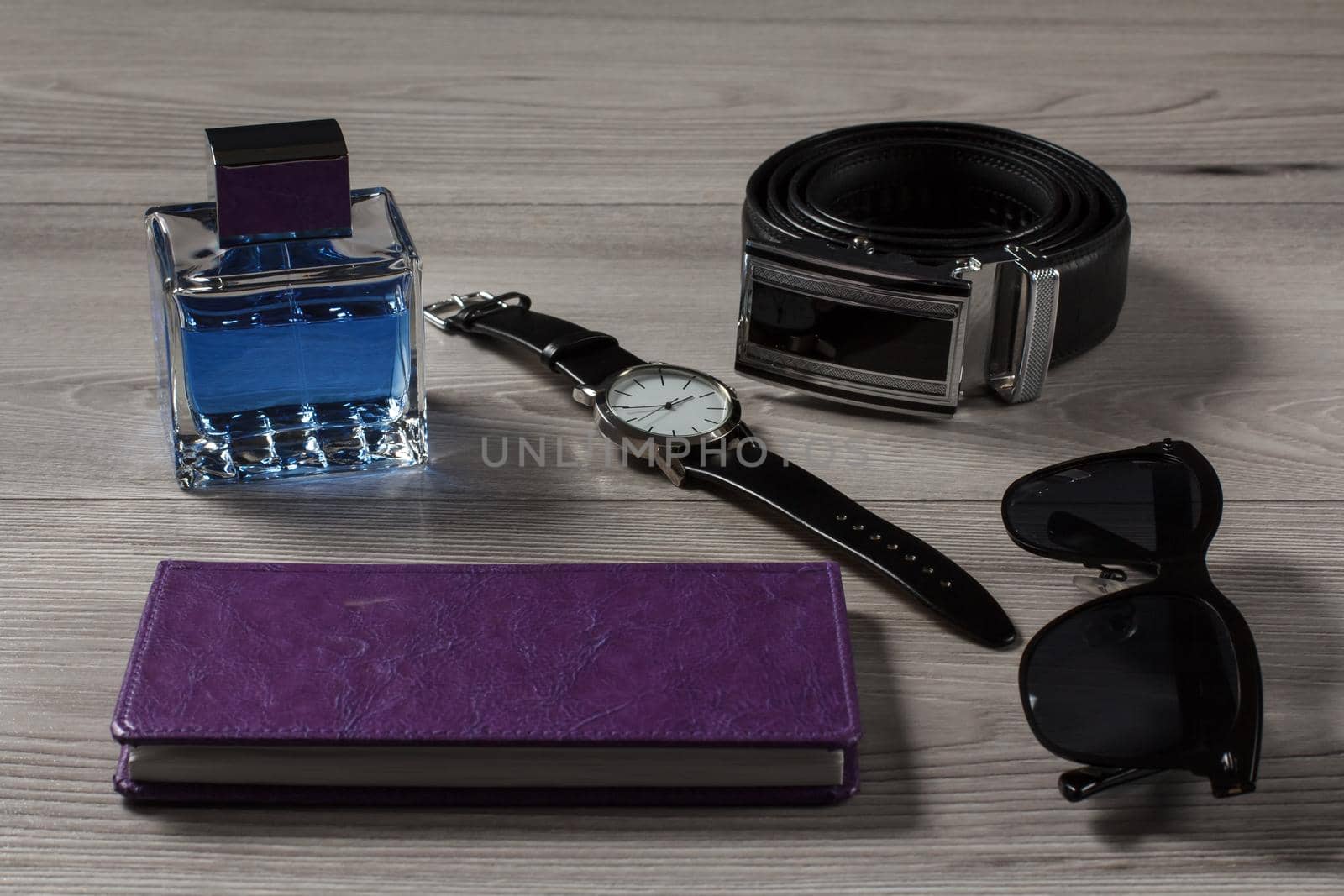 Man perfume, watch with a leather strap, leather belt with metal buckle, notebook in purple cover, black sunglasses on a gray wooden background