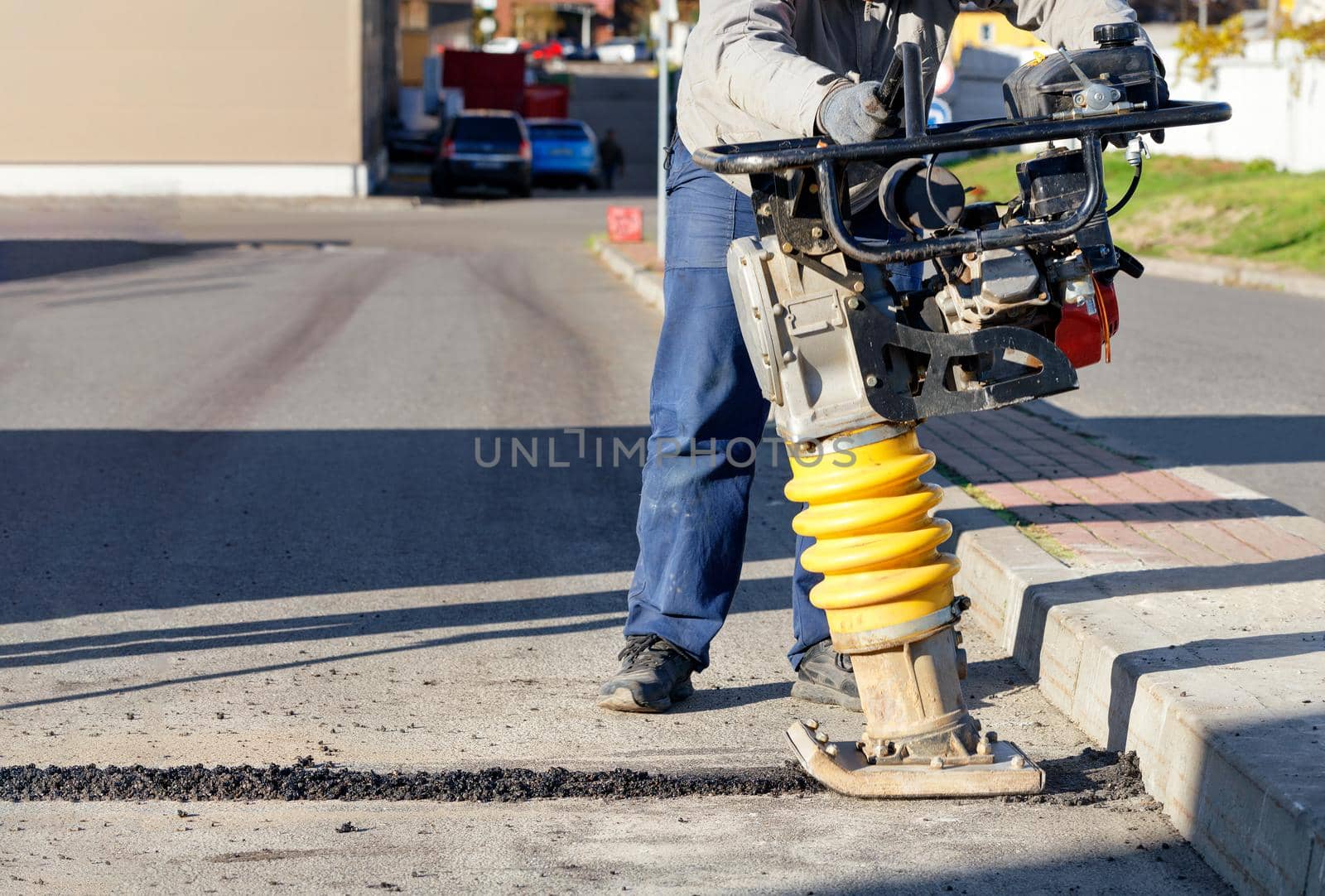 A road worker uses a portable petrol vibration rammer to repair asphalt on the driveway on a bright sunny day.