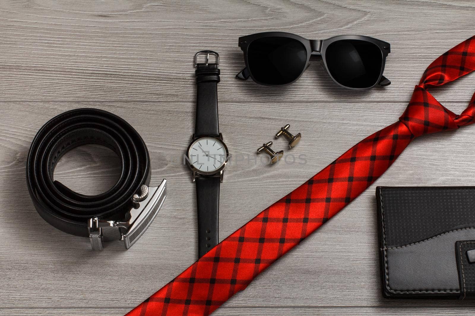 Leather belt with metal buckle, watch with a leather strap, sunglasses, red silk tie, cufflinks, notebook in leather cover on a gray wooden background