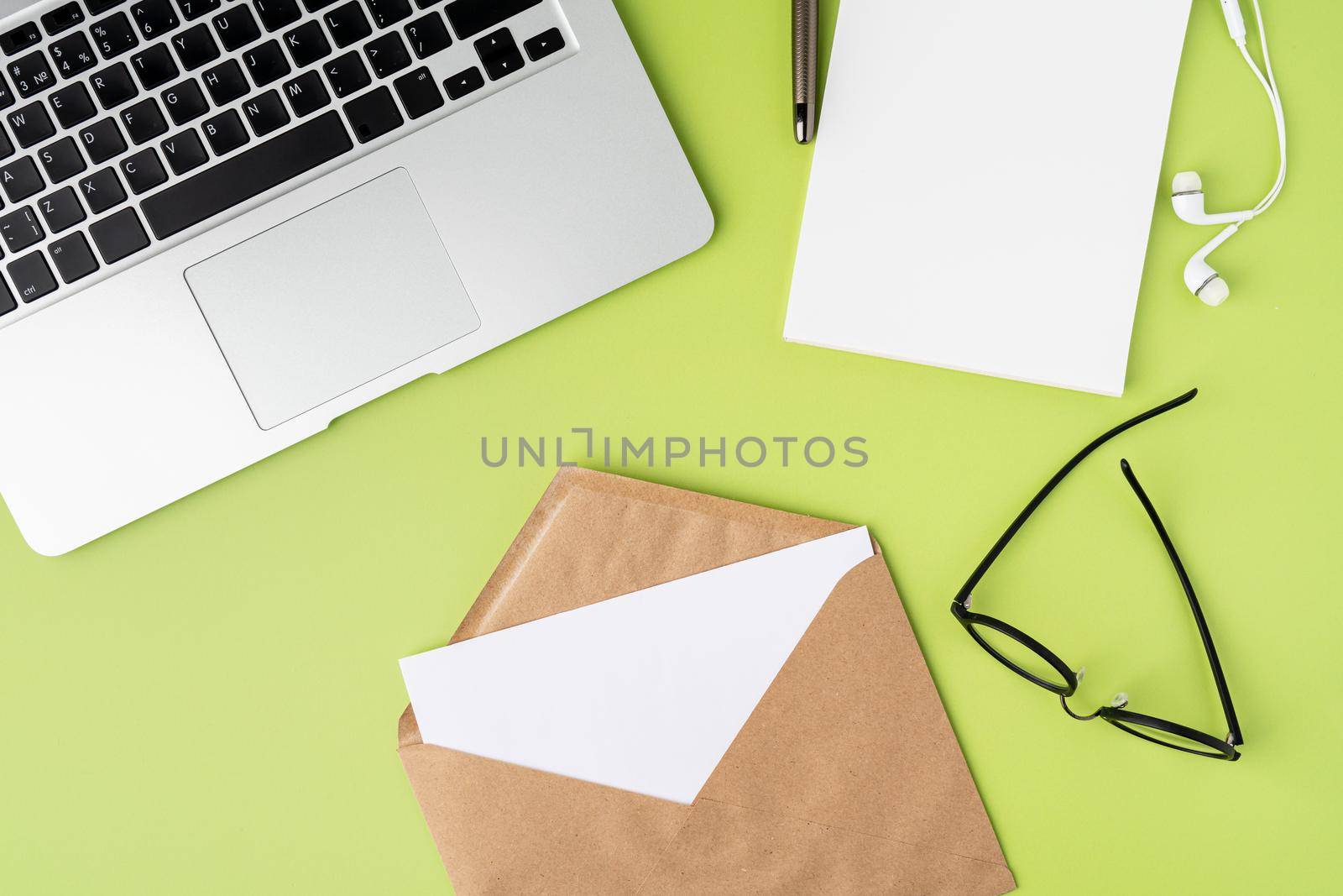 Blank paper sheet in craft envelope on green background. Metallic laptop keyboard, empty notebook, white earphones, glasses, pen. Horizontal from above, copy space