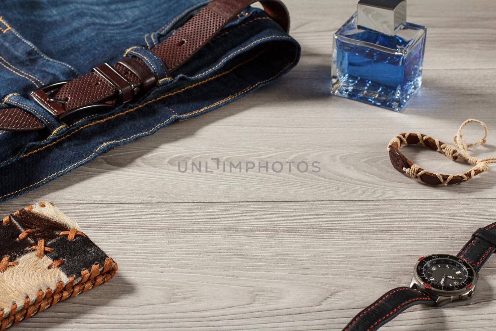 Man perfume, watch with a leather strap, blue jeans with leather belt, amulet and leather purse on a gray wooden background