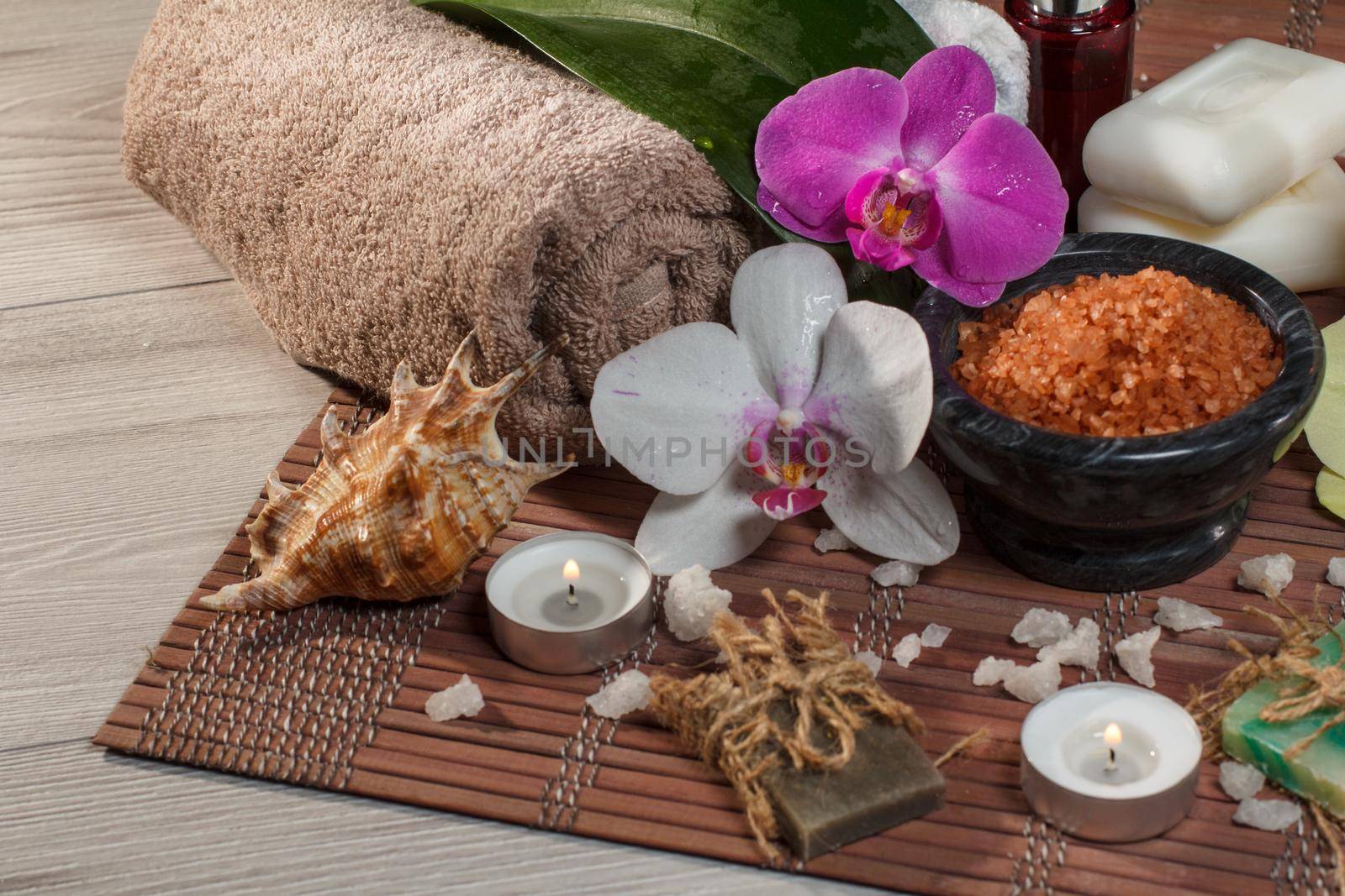 Spa and wellness setting with orchid flowers, bowl with sea salt, seashell, bottle with aromatic oil, soap, candles and towels on bamboo napkin