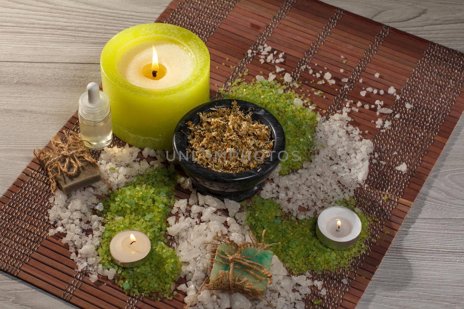 Spa accessories with soap, bowl with dried chamomile flowers, bottle with aromatic oil, green and white sea salt, candles on bamboo napkin