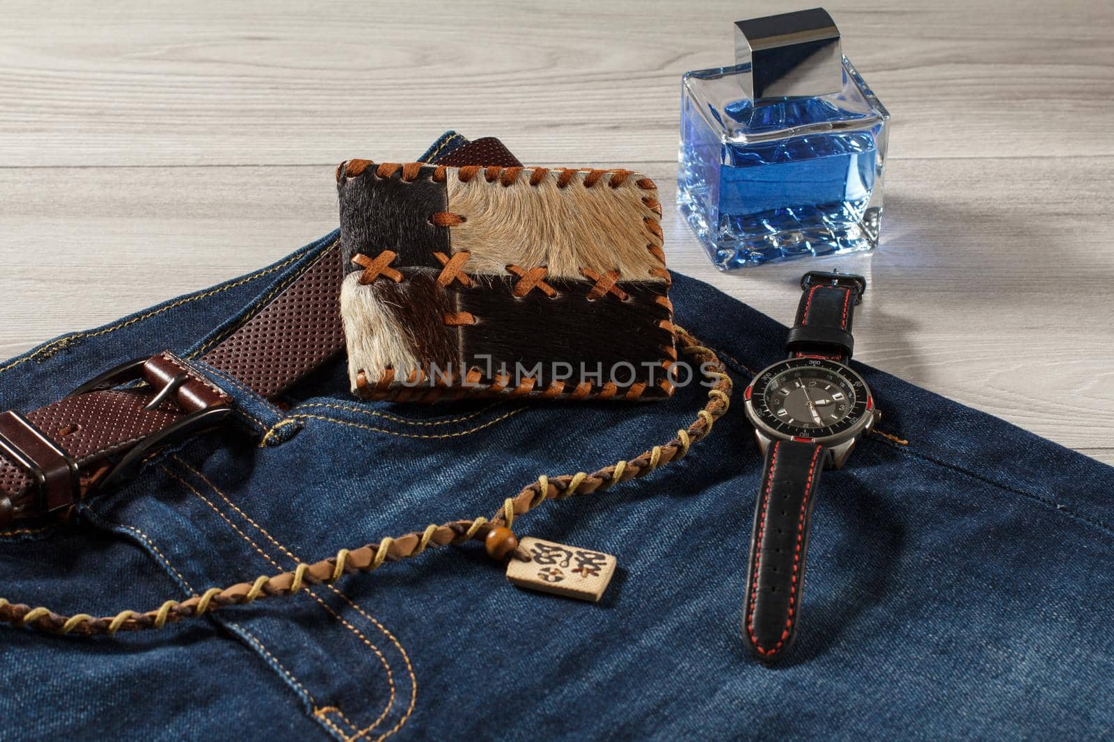 Man perfume, watch with a leather strap, jeans with leather belt, leather purse and amulet on a gray wooden background