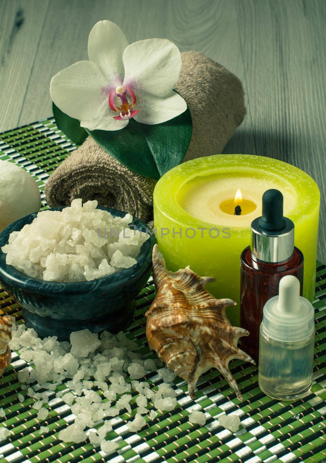 Spa and wellness setting with orchid flower, bowl with sea salt, seashells, bottles with aromatic oil, candle and towel on bamboo napkin and wooden background