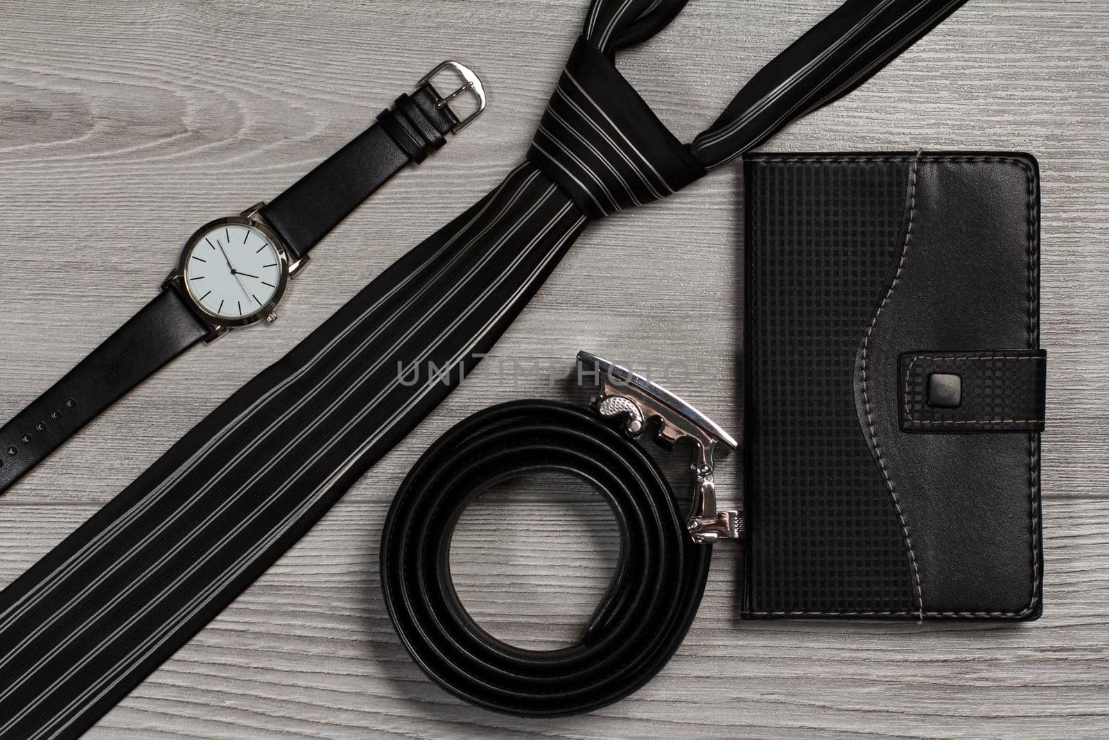 Leather belt with metal buckle, watch with a leather strap, silk tie, notebook in leather cover on a gray wooden background