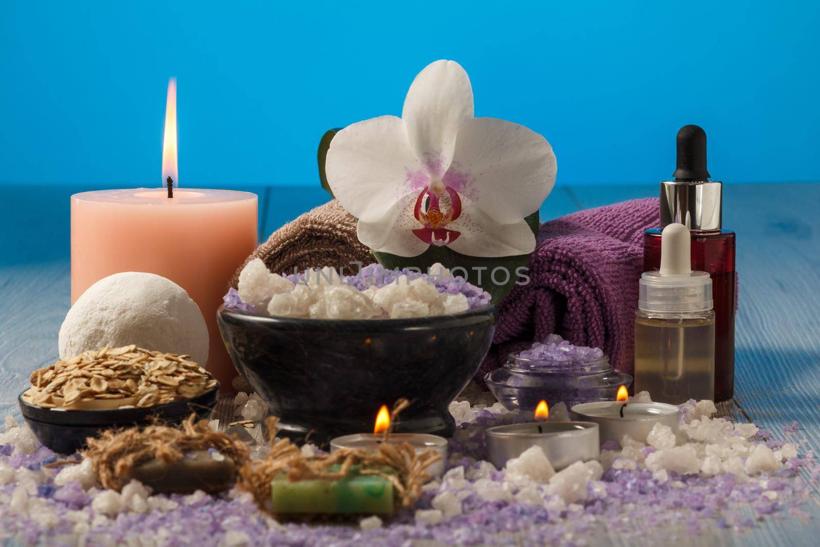 Spa composition with orchid flower, bowl with sea salt, bottles with aromatic oil, soap, scrub, candles and towels on wooden board and blue background