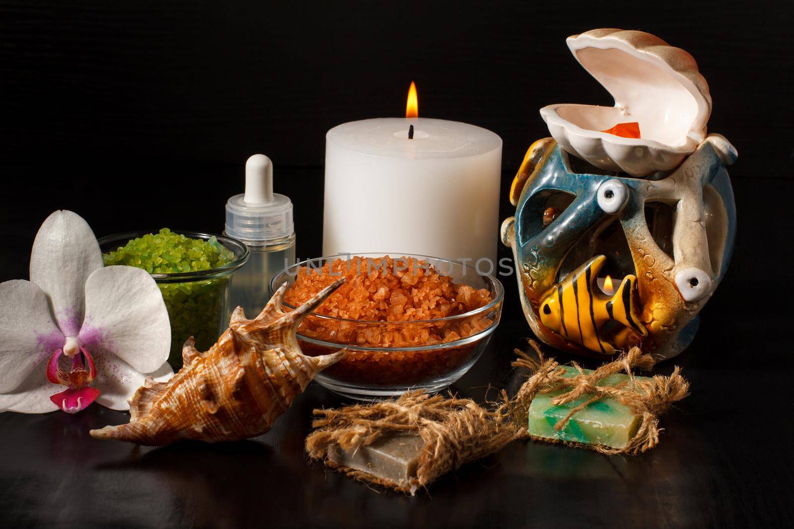 Spa products with orchid flower, handmade soap, bowls with sea salt, bottle with aromatic oil, candles and shell on black background