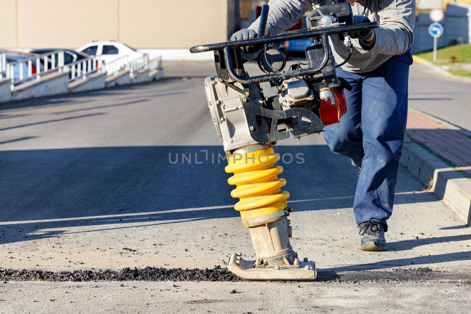 A road worker operates a vibratory rammer to asphalt a small area on a bright sunny day. Copy space, selective focus.