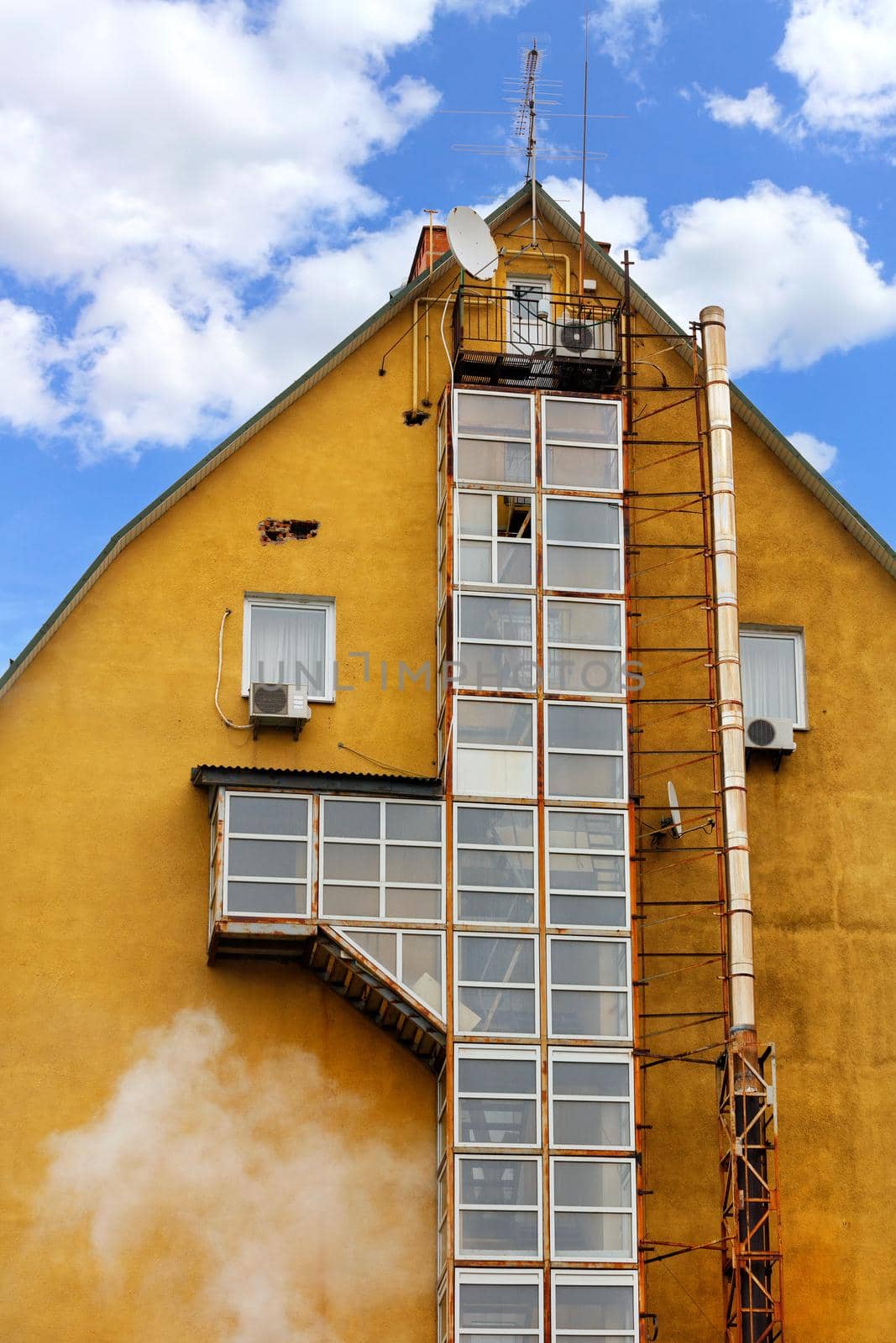 The architecture of an old house, at the butt of which there is a glazed fire escape and a boiler exhaust pipe, external air conditioners and a satellite dish against a blue sky with clouds.