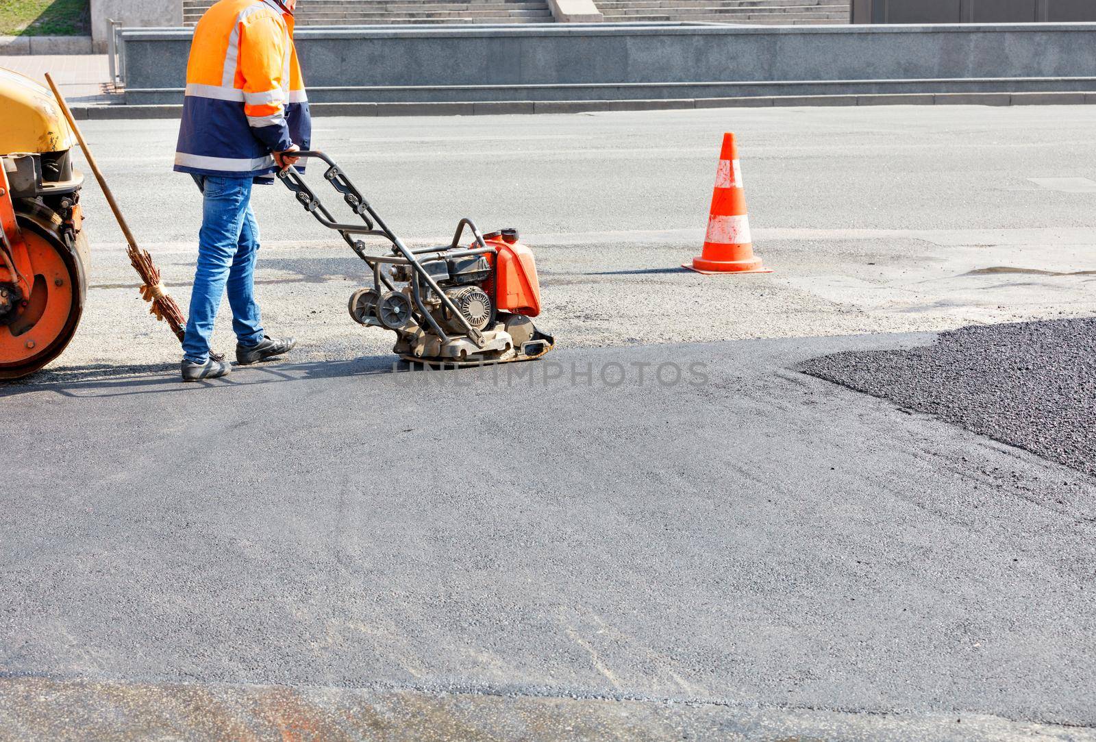 A road worker compacts the asphalt with a petrol vibrator on a fenced road. by Sergii