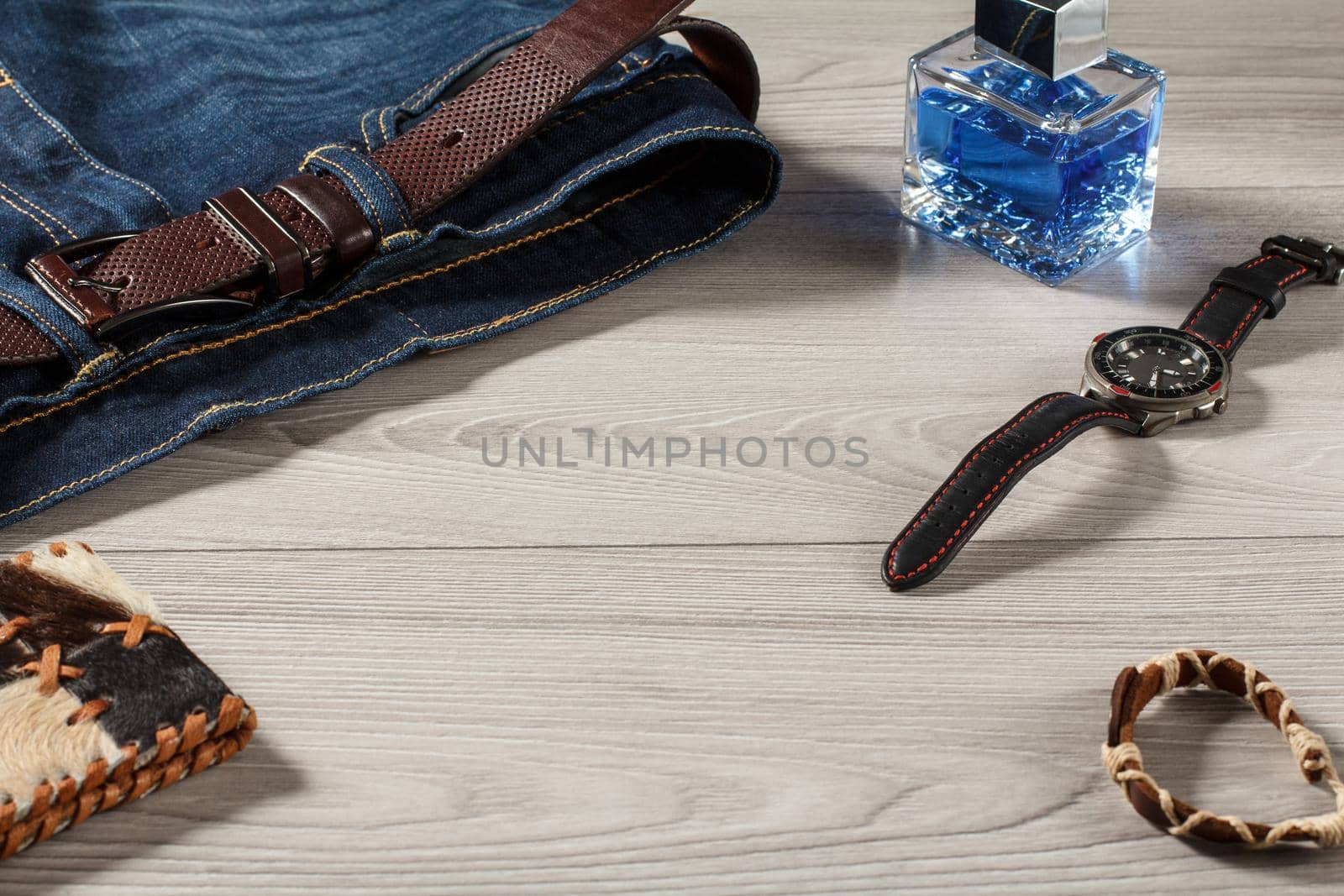 Man perfume, watch with a leather strap, jeans with leather belt, amulet and leather purse on a gray wooden background