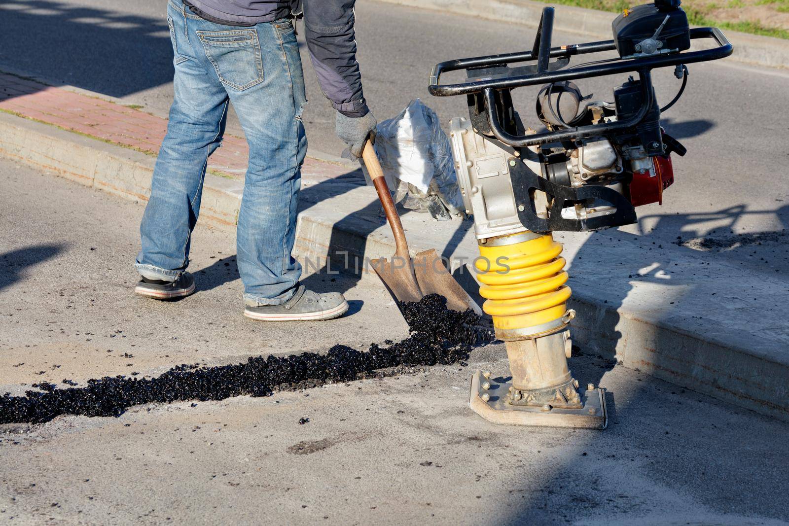 Cold asphalt is used by road workers to repair and seal cracks on the roadway on a bright sunny day, against the backdrop of a rammer.