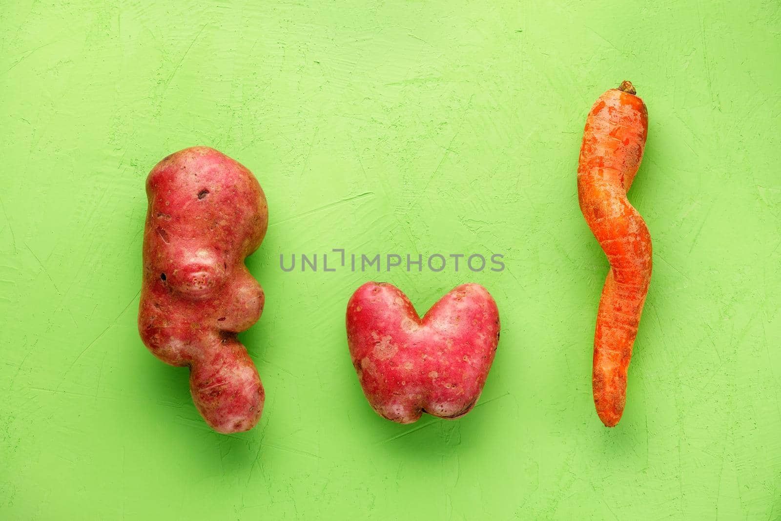 Ugly potatoes and carrots on a background of green plaster. Vegetable or food waste concept. Top view, close-up. by Sergii