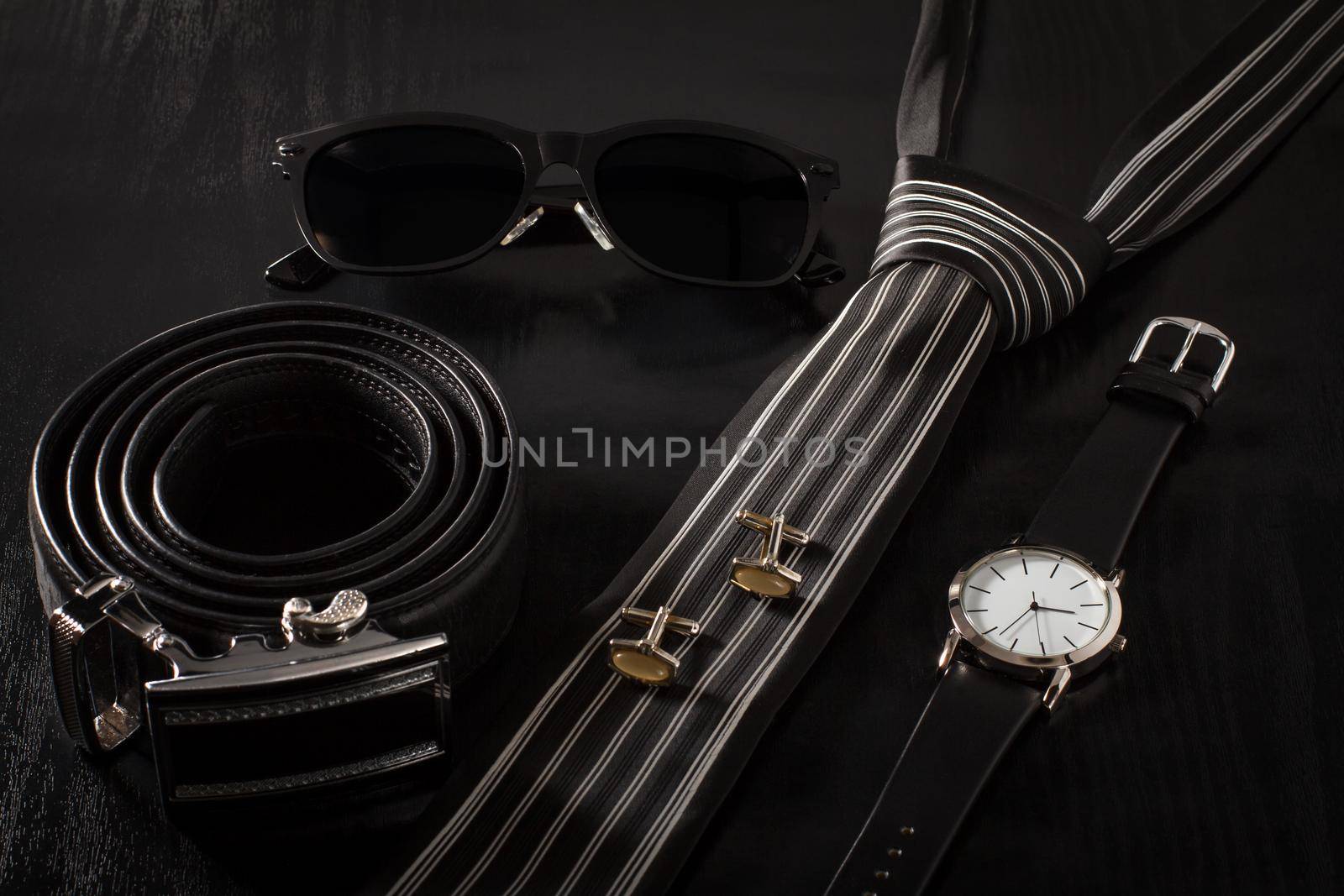 Leather belt with metal buckle, sunglasses, tie, cuff-links, watch with a leather strap on a black background