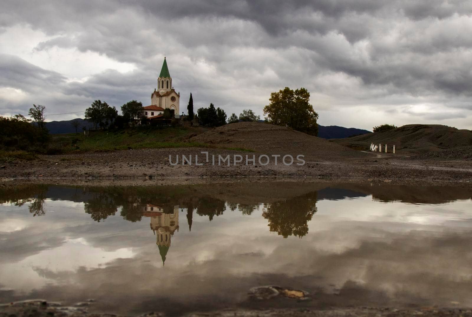 Landscape showing Puig Agut church and some trees under a cloudy sky and its reflection on a puddle