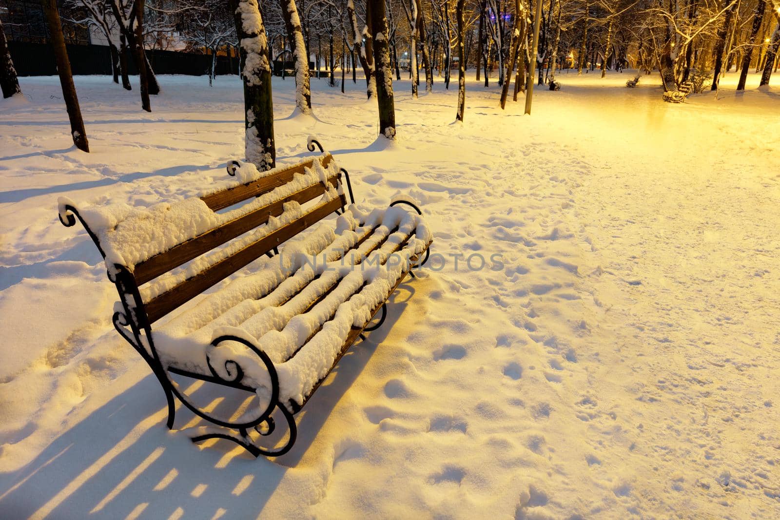 Empty brown wooden bench with metal decorative railing in the snow under the warm light of a street lamp on a winter evening in a city park.