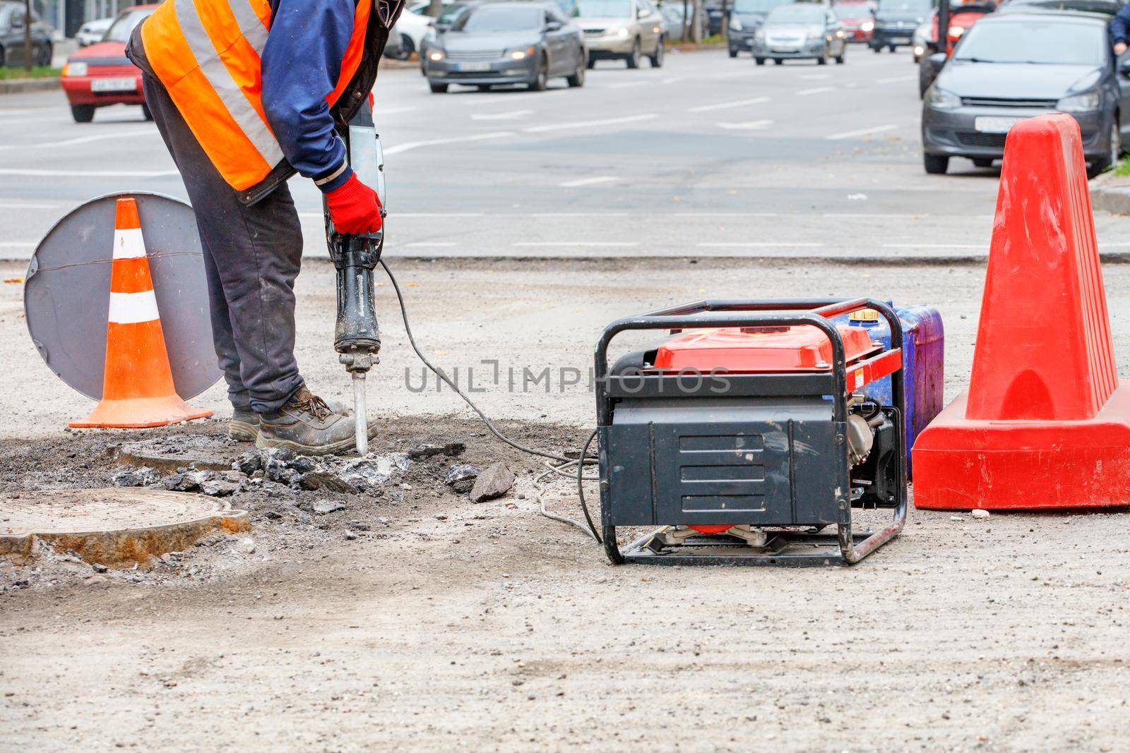A road worker in reflective clothing breaks old asphalt off the road with an electric jackhammer. by Sergii