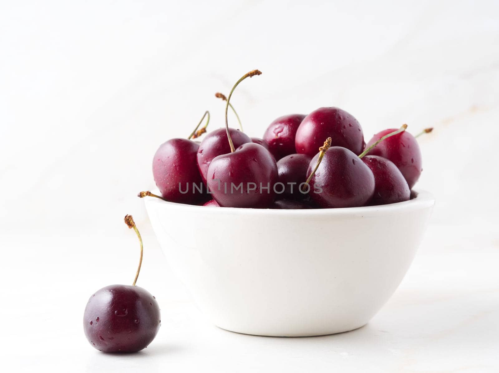 The red dark sweet cherries in white bowl on stone white table, side view.