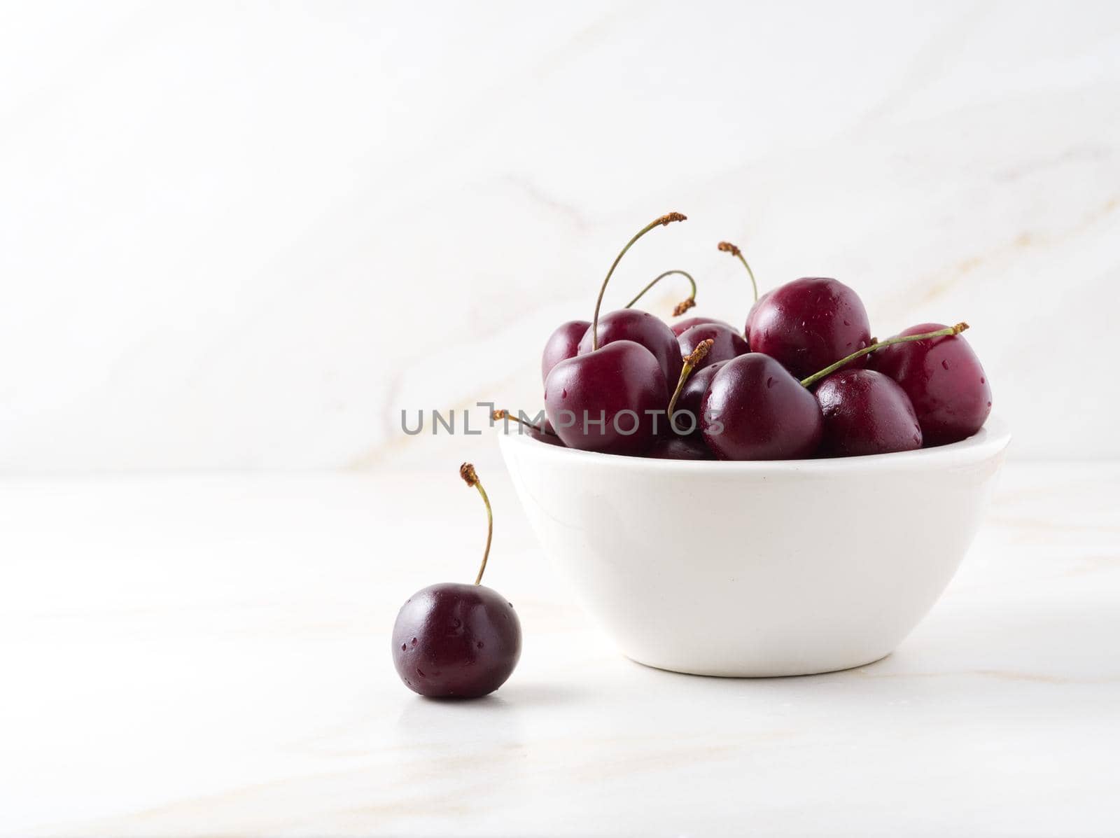The red dark sweet cherries in white bowl on stone white table, side view, copy space