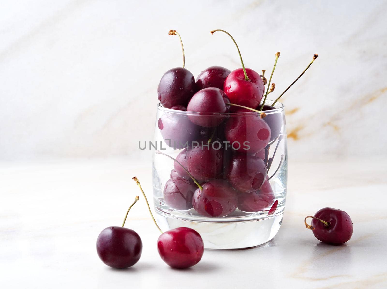 The red dark sweet cherries in glass on stone white table, side view, copy space