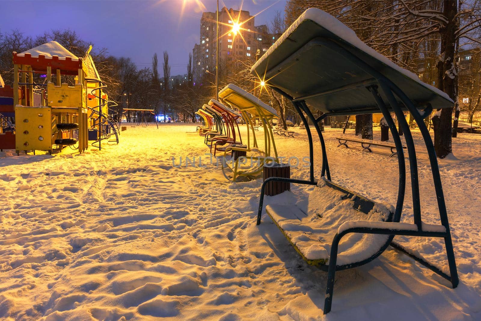 Wooden benches and a playground in the winter city evening park are covered with snow against the background of blue twilight. by Sergii