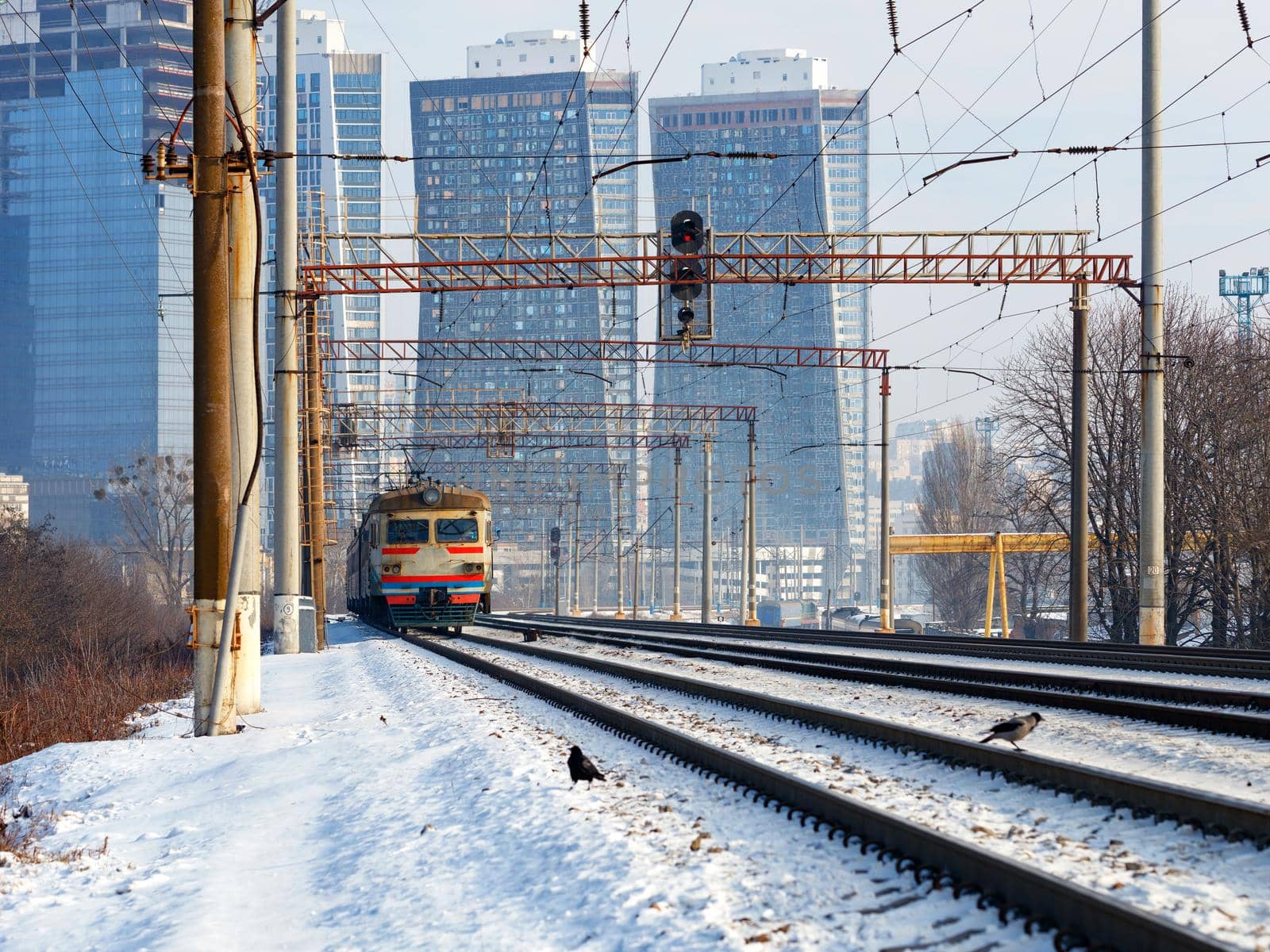 An old electric train moves on rails in the winter season against the backdrop of a cityscape and skyscrapers under construction in the light of sunlight.