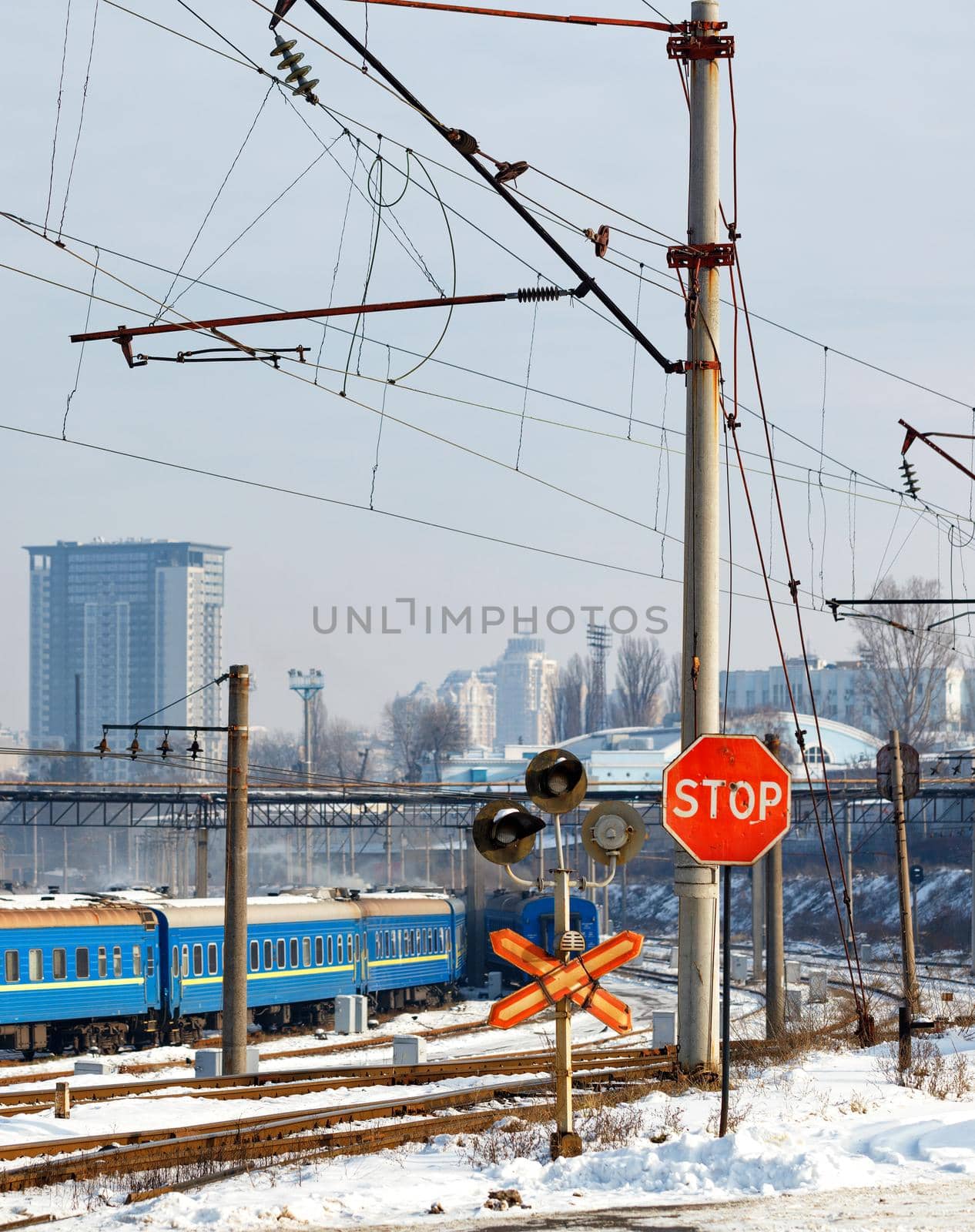 Red stop sign at a railway crossing against the background of railway tracks, blue train carriages and cityscape at the entrance to the city, winter cityscape, vertical image.
