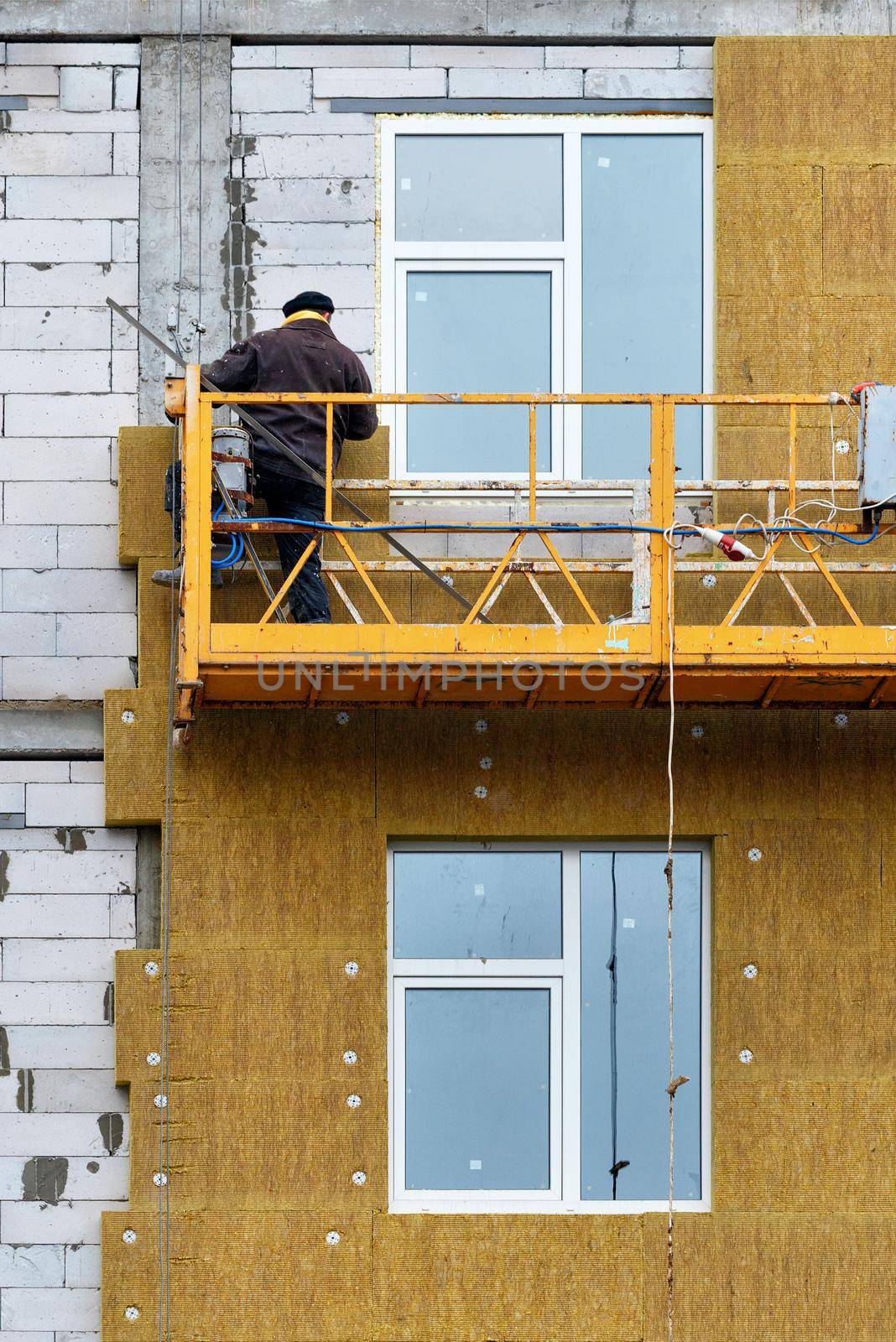 The builder stands on a suspended platform and insulates the facade of a house under construction. by Sergii