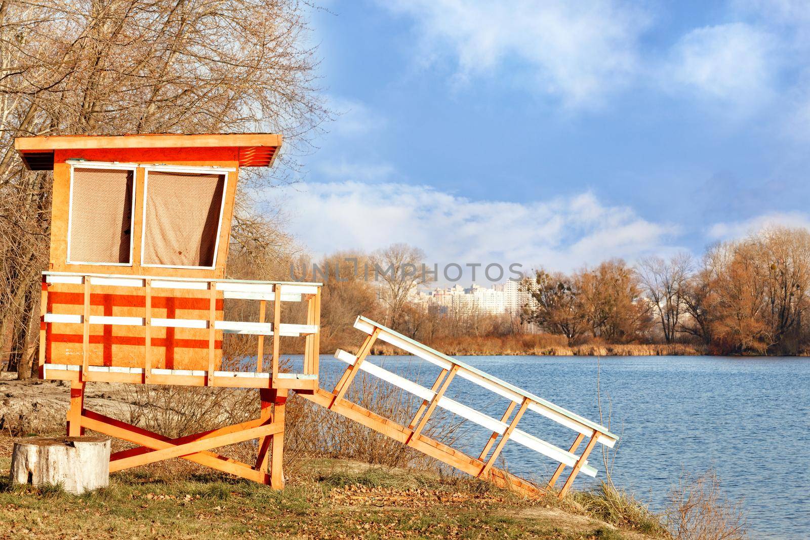 On the bank of the river with bare autumn bushes without leaves stands a lonely wooden house of lifeguards and is illuminated by the rays of the bright autumn sun. by Sergii