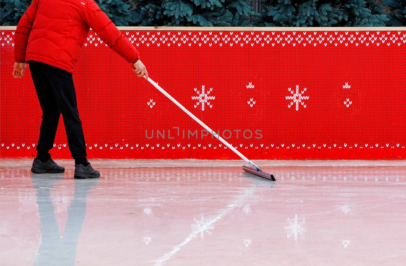 An ice stadium worker, surrounded by a red fence and wearing a red jacket, scrubbing the ice surface with a rubberized mop to remove small chips. Copy space.