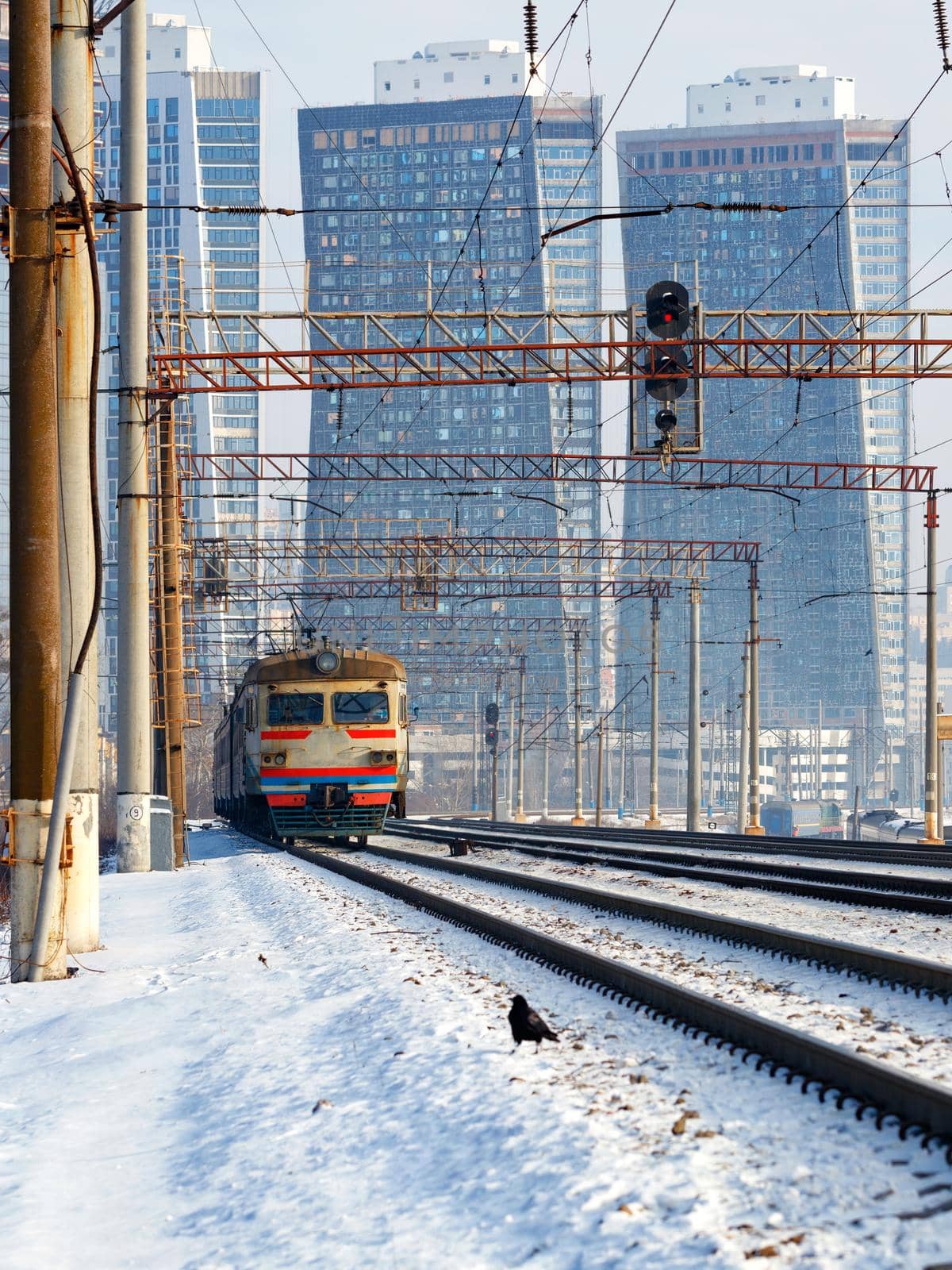 An electric train moves on rails against the backdrop of a cityscape in a winter haze, vertical image.