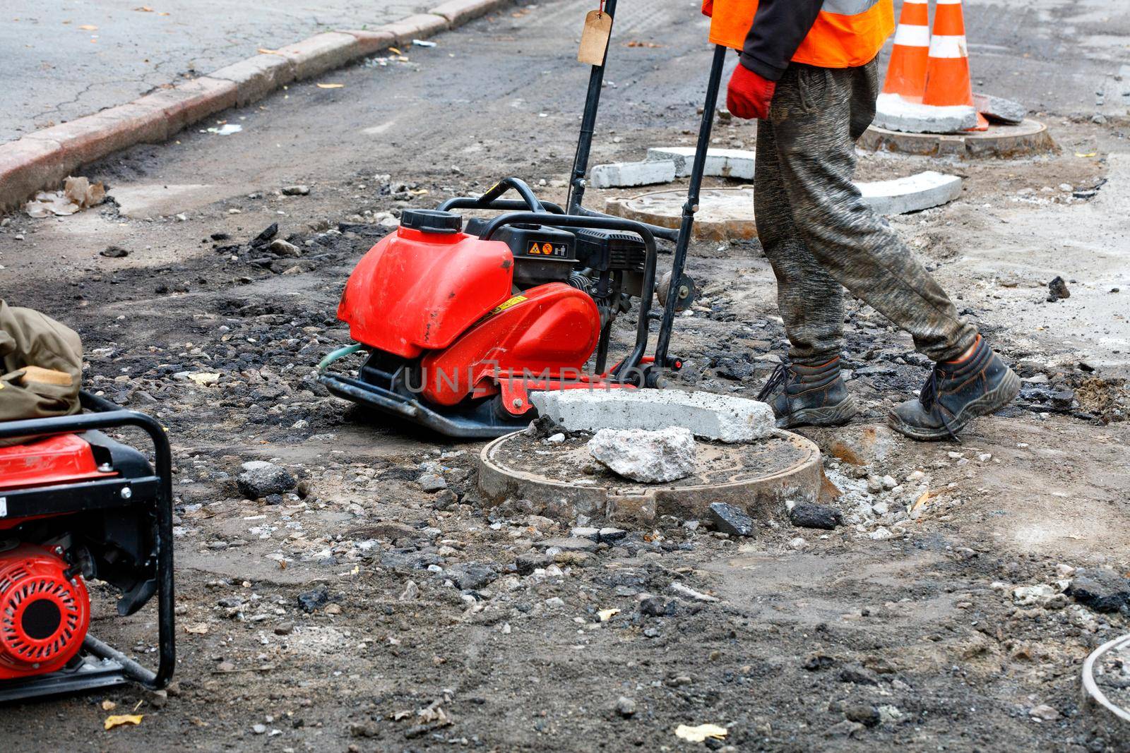 A road worker wearing a reflective orange vest repairs and installs sewers using a compactor plate, an electric jackhammer and a petrol generator on a roadway surrounded by red cones. Copy space.