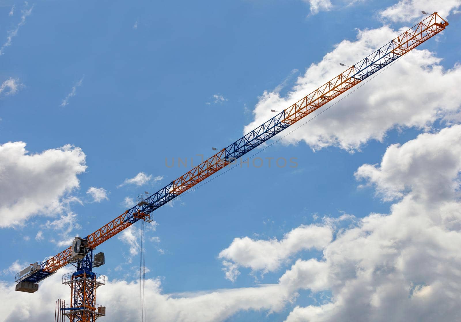 The arrow of a construction tower crane in yellow and blue is located diagonally from bottom to top against a background of blue sky and white clouds, copy space.