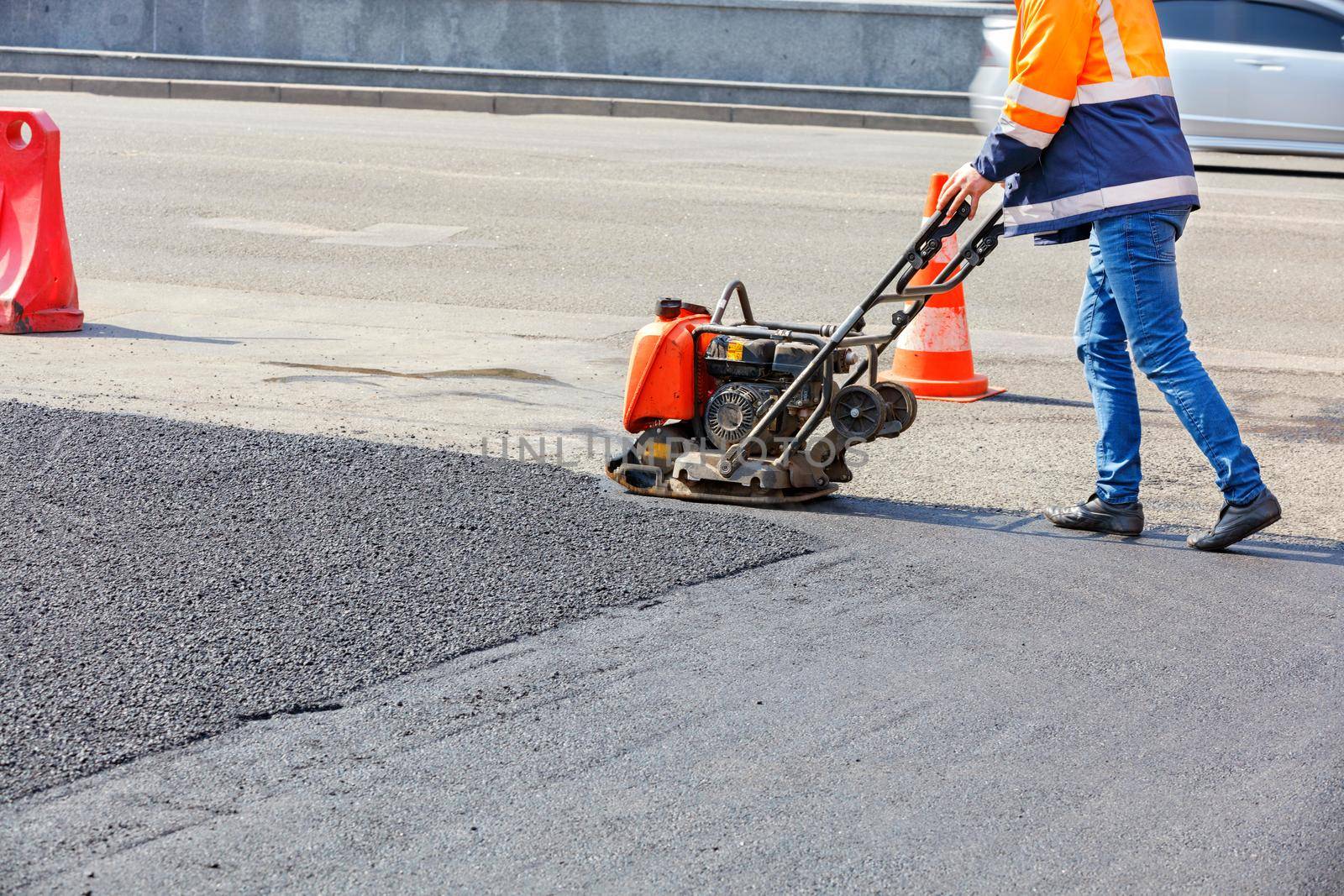A road worker uses a vibratory plate to compact asphalt on a fenced road repair site on a bright sunny day.