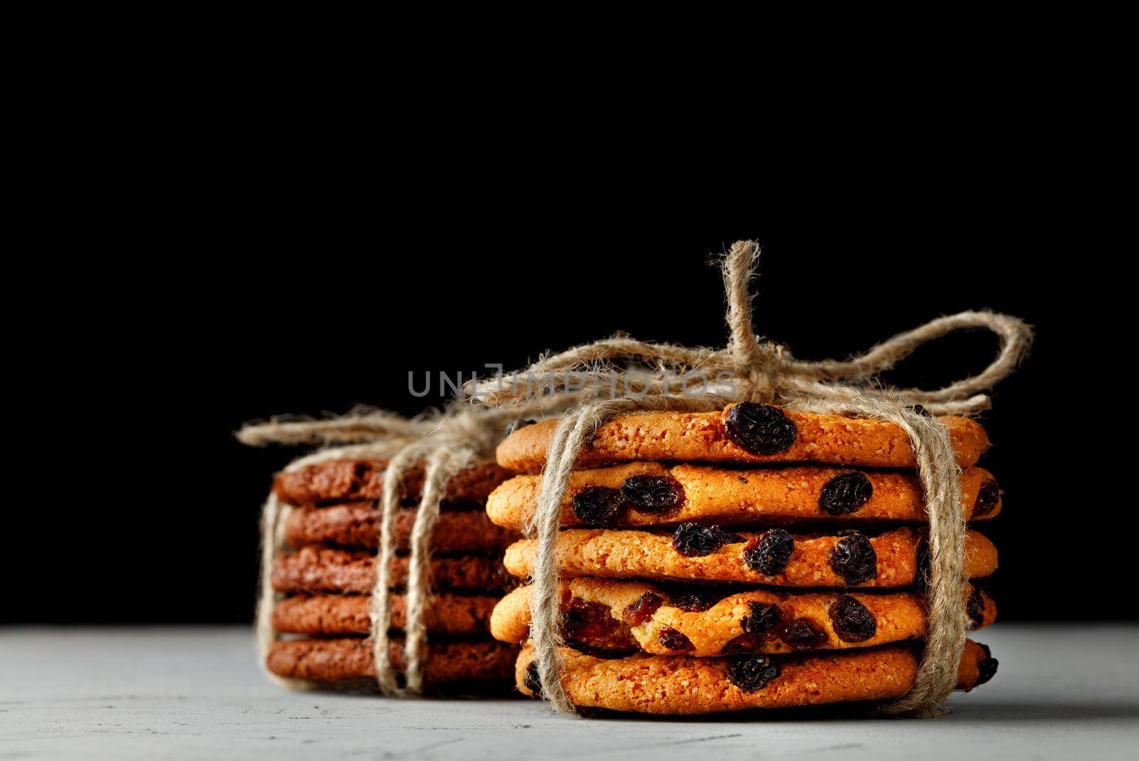 Stacks of homemade raisin oatmeal cookies and dark chocolate biscuits tied with rope against a black background. Copy space. Close-up. Selective focus.