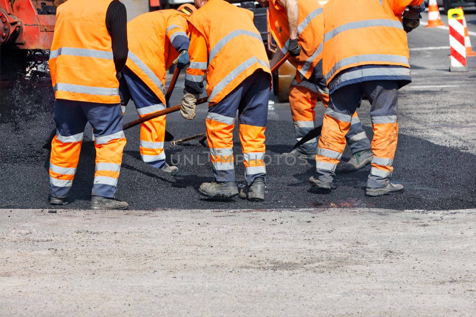 A team of road workers in orange reflective uniforms cover a section of road with fresh asphalt to repair the section of the road. Clear day, copy space.