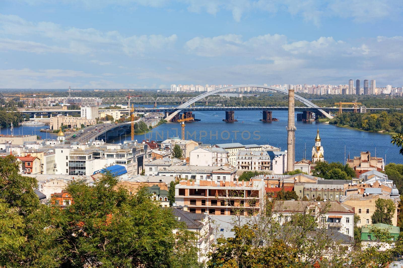 A view of the old Podil district of summer Kyiv, automobile and railway bridges, a bell tower with a gilded dome, the Dnipro River and many old and new city buildings.