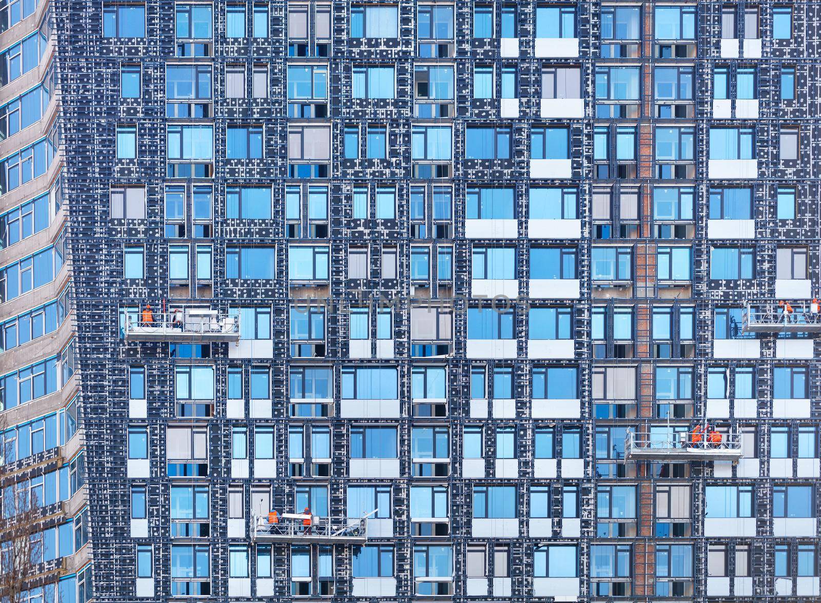 Several teams on construction cradles are insulating a modern house under construction, the background and texture of glass windows, a fragment of the facade with a reflection of the blue sky.